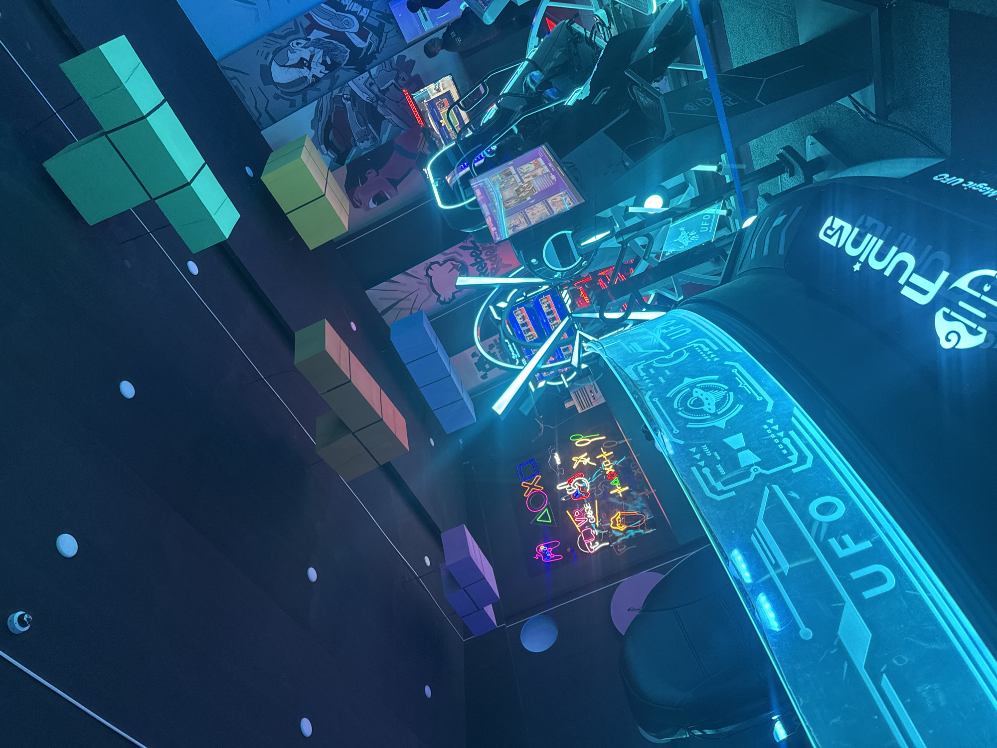 The image captures an interior scene within FuninVR Magic UFO in Abuja, Nigeria. The perspective is from a low angle, looking upwards towards a ceiling adorned with neon lights and geometric shapes resembling Tetris blocks. The walls are a dark blue or black. Various neon signs are visible, including a game console icon display and a large “UFO” sign. A black gaming chair is partially visible in the foreground. The scene has a modern, playful, and futuristic aesthetic.