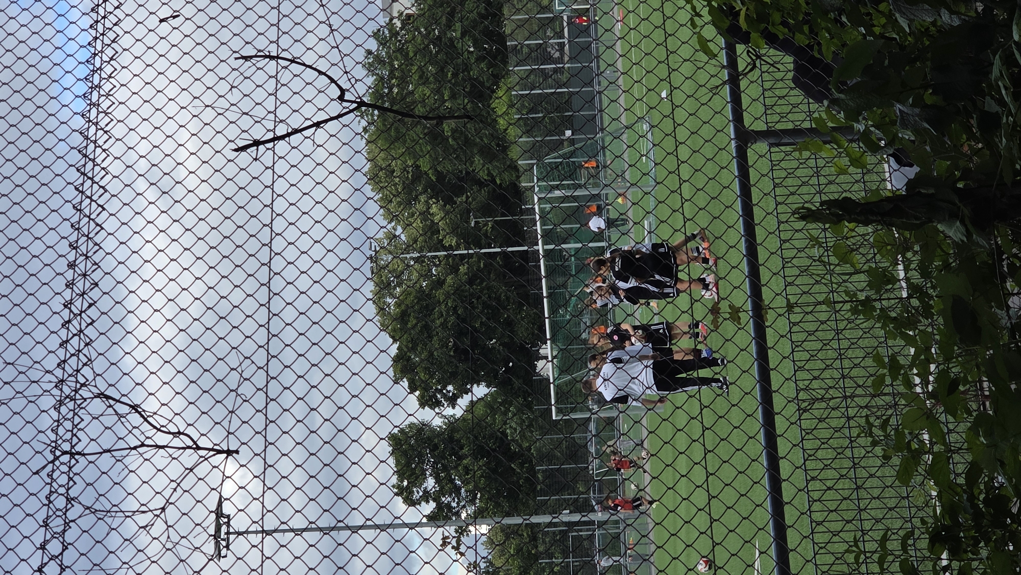 The image shows a soccer field through a chain-link fence with barbed wire at the top. There are several people, mostly women, in athletic wear on the field, appearing to be playing or practicing soccer. Some are wearing black and white uniforms. There are soccer balls visible, and a goal net is in the background. Trees and buildings are visible beyond the field. The sky is overcast.