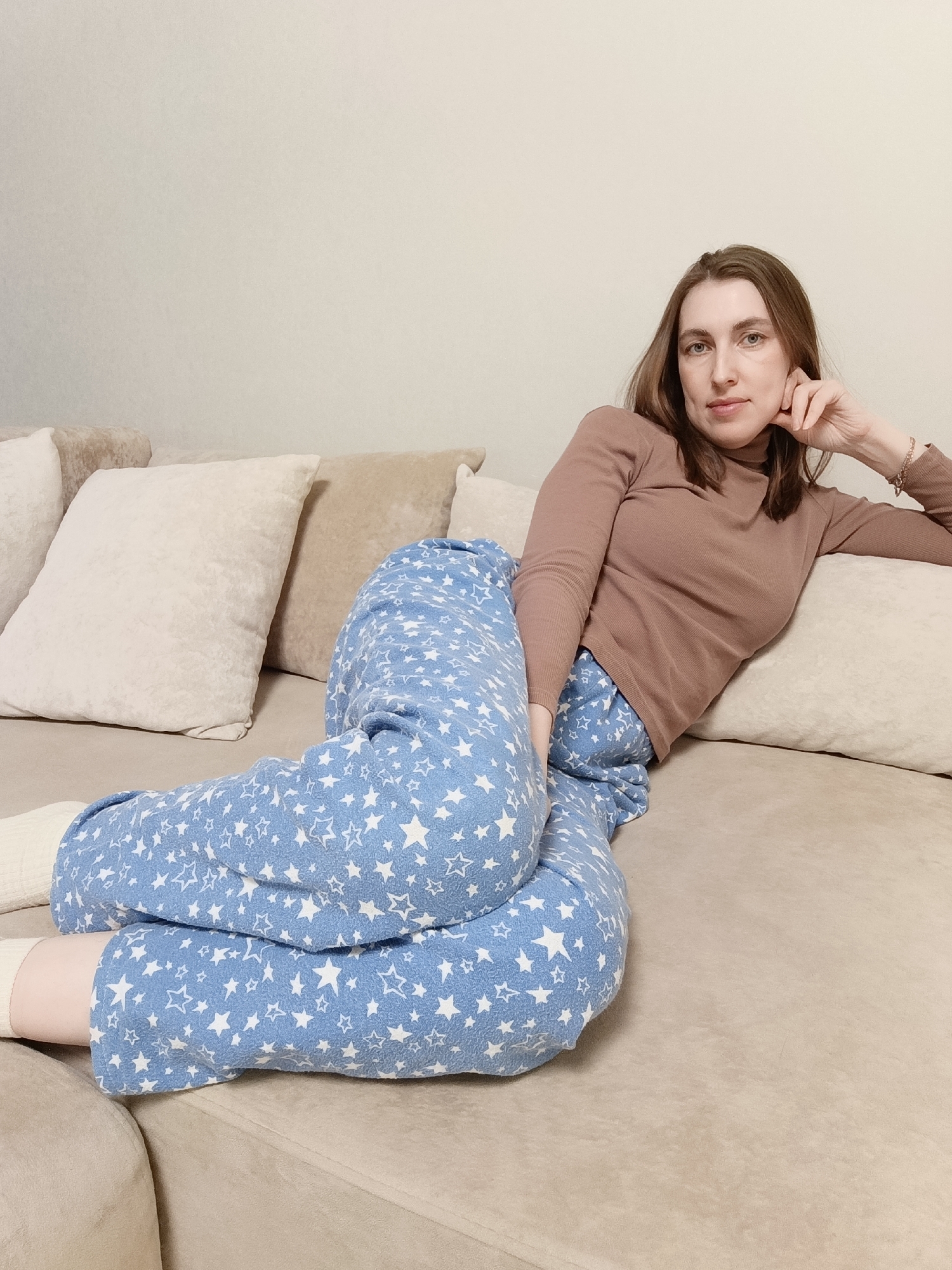 A woman is reclining on a tan-colored sofa. She is wearing blue pajamas with a white star pattern and a brown long-sleeved top. She has her head resting on her hand and is looking toward the camera. Behind her are several pillows in beige tones. The background is a light beige wall. The image was taken inside. There is no information available in this image regarding the location of this photo as Chaikovsky, Russia.