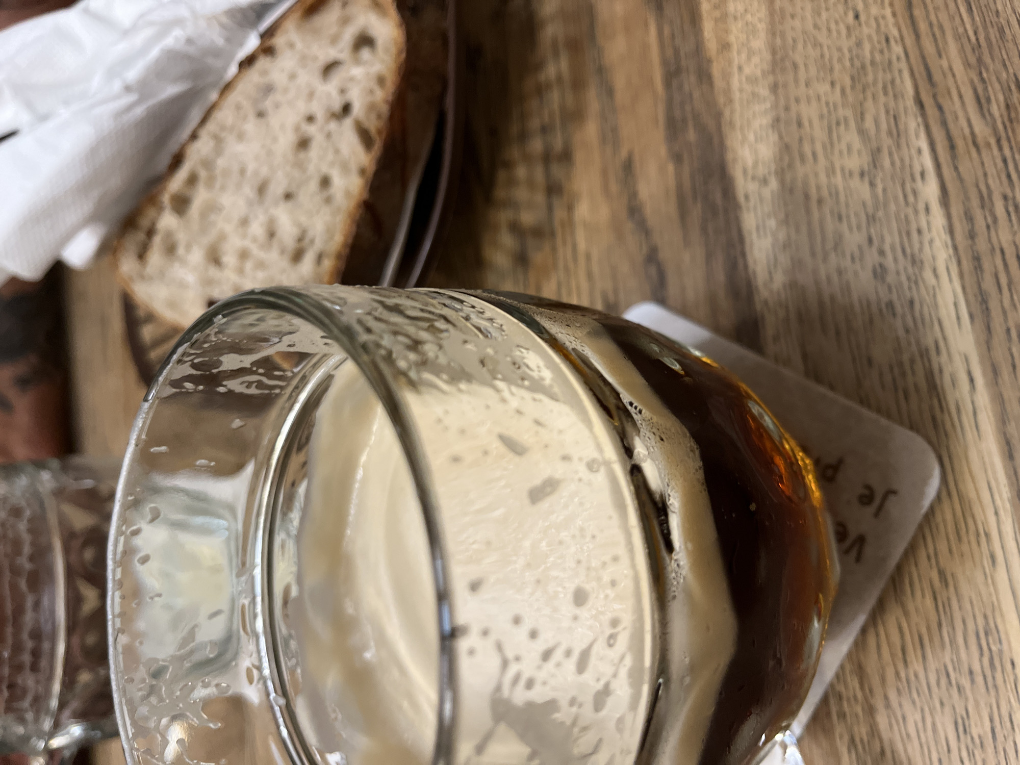 A close-up, slightly elevated shot captures a half-full glass of dark amber liquid topped with a substantial layer of creamy beige foam, positioned on a light-colored coaster. The glass displays condensation and foam residue on its inner surface, with the liquid level at approximately one-third of the glass's height. Faint text, reading "Ve" and "Je P," is visible on the coaster beneath the glass. In the blurred background, to the right, a single slice of rustic-style bread with an irregular, open crumb structure rests on a dark plate. A crumpled white napkin is situated above the bread. The entire scene is set on a warm-toned wooden table, showing distinct grain patterns. The lighting suggests an indoor setting, likely a cafe or restaurant in Unknown, Unknown. No people are visible.