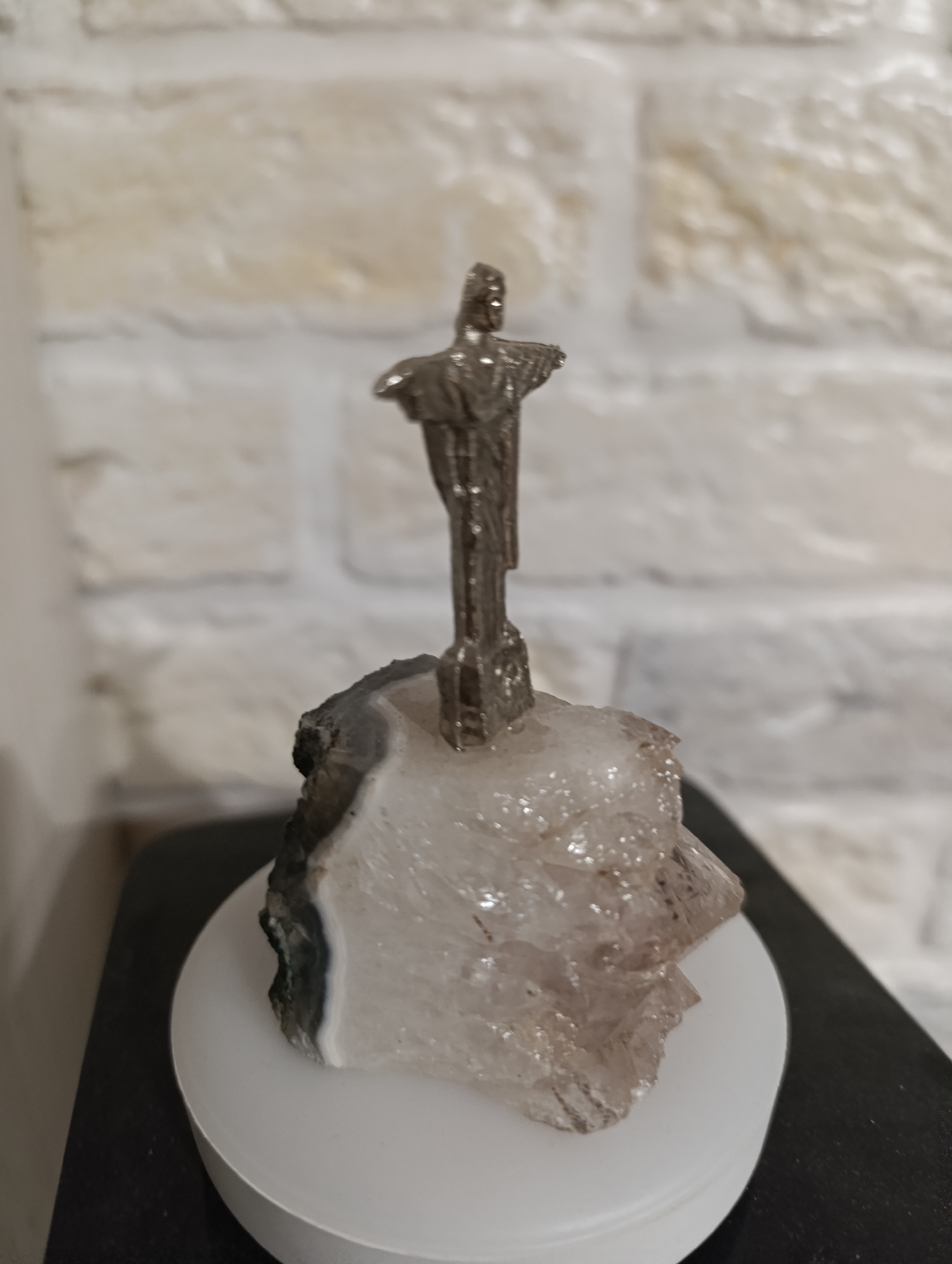 A small, silver-toned figurine of Christ the Redeemer stands atop a rough, translucent mineral specimen. The figurine, depicting Christ with arms outstretched, is a miniature replica. The mineral, likely quartz or a similar crystalline formation, has layers of white and grey, with some darker, more opaque inclusions. The mineral rests on a smooth, white circular base, which in turn sits on a dark, flat surface. The background consists of a lightly textured, light-colored wall, possibly made of bricks or stone. The lighting is soft, casting a gentle illumination on the objects. The scene appears to be an indoor display or decorative item. The image is taken from a slightly low angle, focusing on the figurine and its base. There is no indication of location within a city.