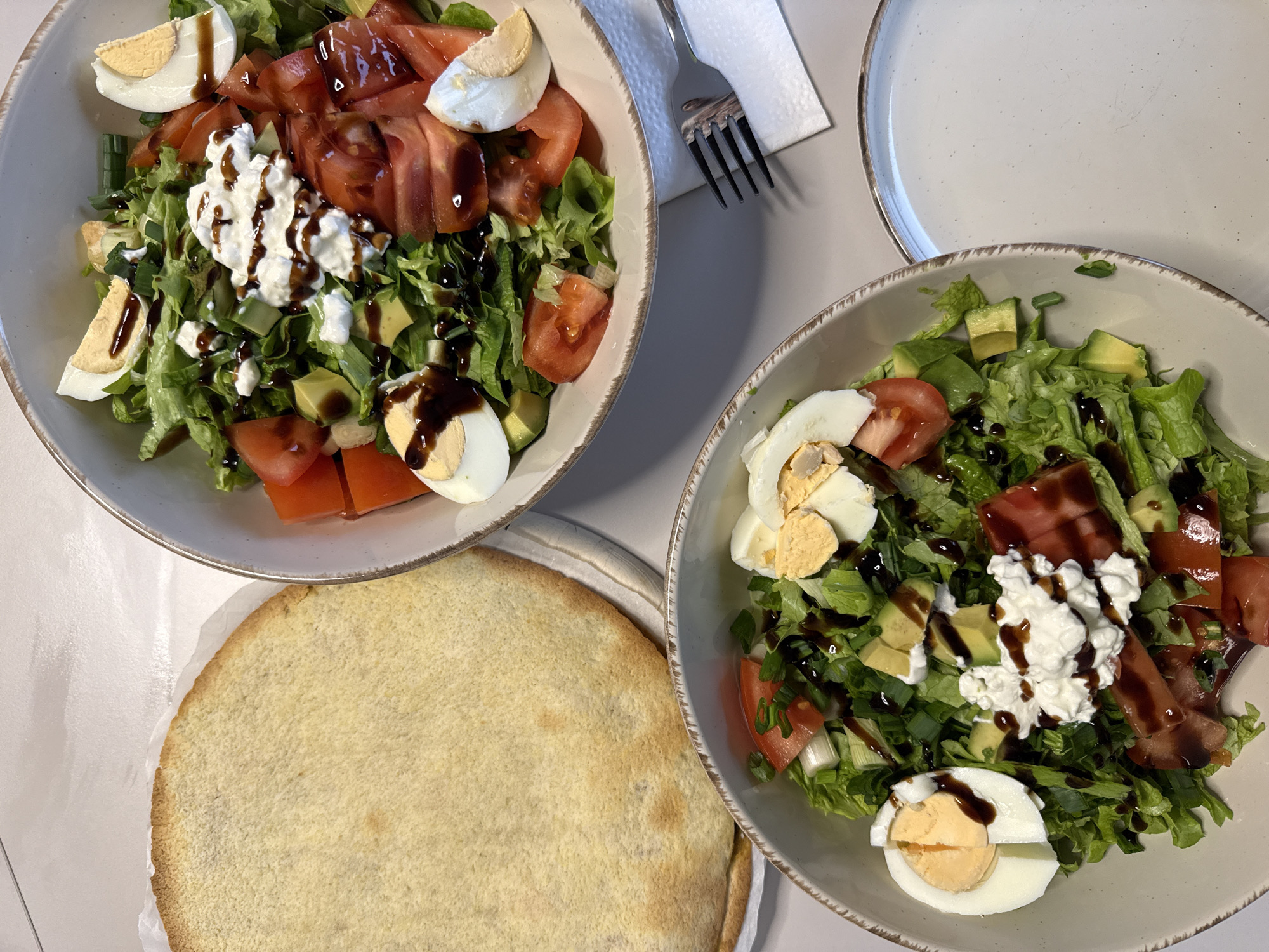 The image shows a tabletop setting, likely in a home, with two bowls of salads and a round flatbread. Each salad bowl contains lettuce, tomatoes, avocado, hard-boiled eggs, and a dollop of white cheese, drizzled with balsamic glaze. Next to one of the salad bowls is a fork and a napkin. A plate is also visible. The flatbread is a plain-looking, round baked item. The location is unknown.