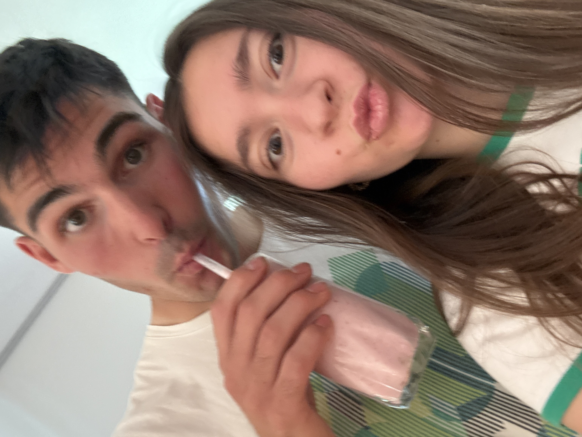 A close-up selfie shows a young man and woman. The man, on the left, is drinking a pink beverage from a clear plastic cup with a straw. He has short dark hair, light skin, and dark eyes. The woman is slightly behind him with her head tilted towards him. She has long brown hair, light skin, and a mole on her cheek. They are both wearing white shirts, with the woman's shirt having a green stripe. The background is blurry and indistinct. There is no clear indication of the location being Sofia, Bulgaria.