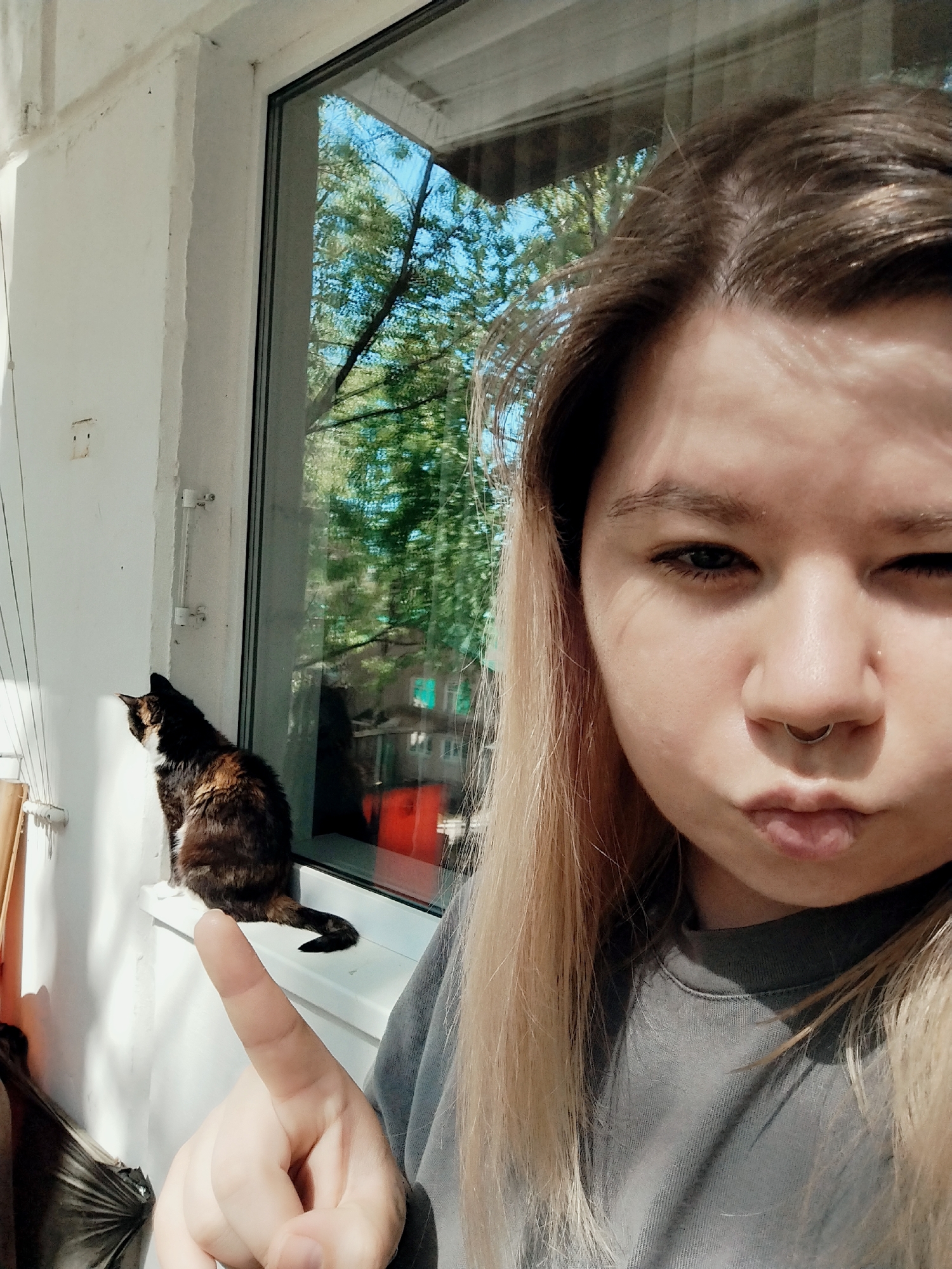 A woman with blonde hair and a septum piercing is taking a selfie on a balcony. She is making a puckered face and pointing with her index finger towards a calico cat sitting on the windowsill. Outside the window, trees and a building can be seen.