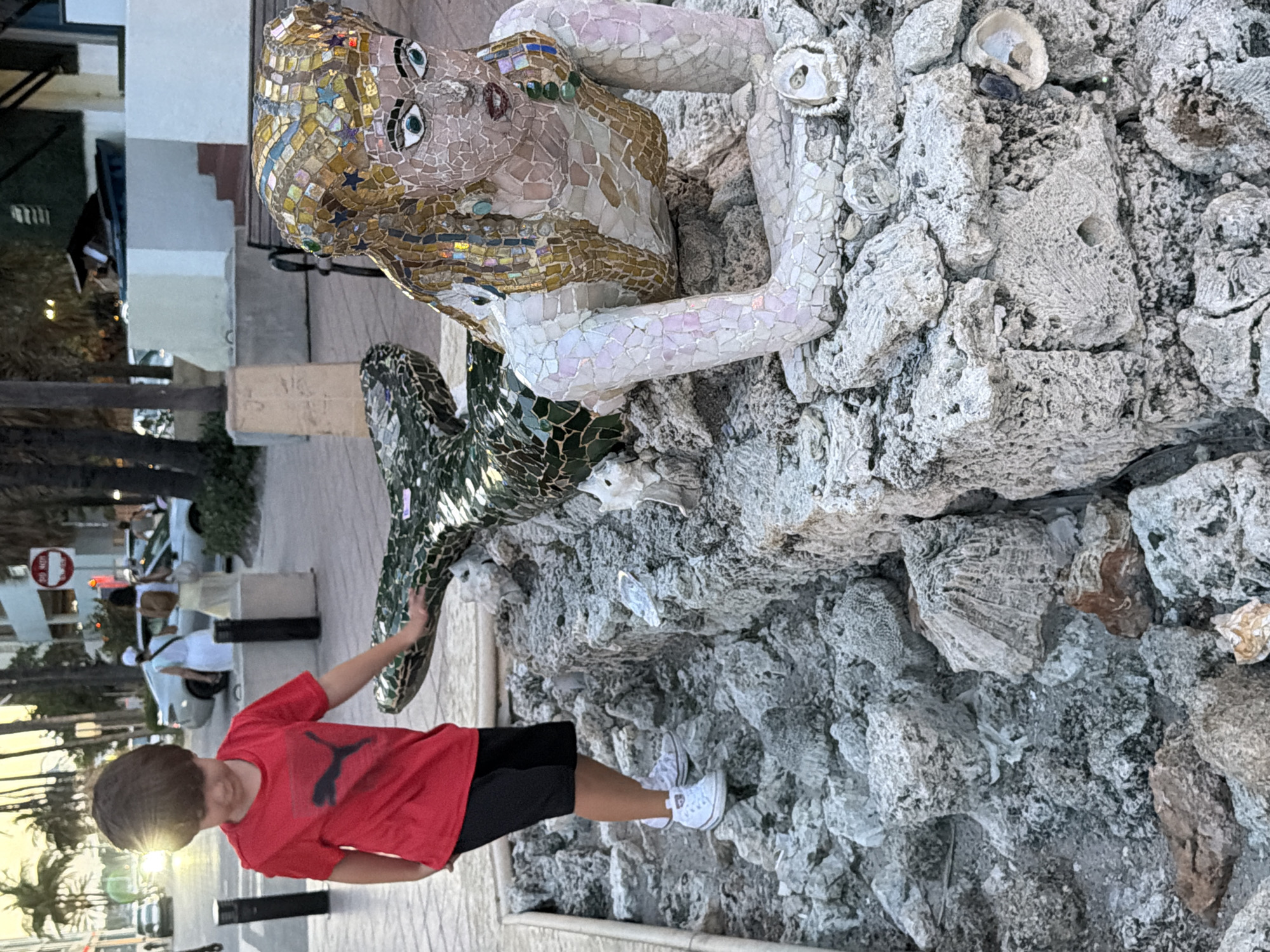 A young boy in a red t-shirt and black shorts stands on a rocky structure, reaching out to touch a mosaic sculpture of a mermaid. The boy is positioned on the left side of the frame, with his right hand extended towards the mermaid's tail. The mermaid sculpture, made of colorful tiles, is lying on its side, with its head facing right. The background shows a street scene with palm trees, buildings, cars, and pedestrians, indicating an outdoor urban environment, likely in Hollywood, United States of America. The lighting suggests it is late afternoon or early evening.