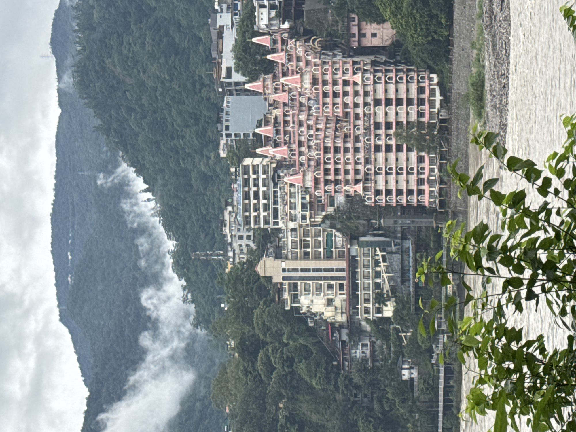 The image captures a view of the Ganges River flowing through Rishikesh, India, with a backdrop of lush green mountains. In the midground, a distinctive pink and white building with multiple spires, resembling a castle or temple, dominates the scene. This building is situated on a hillside, surrounded by other multi-story structures and dense foliage. The river in the foreground has a murky, brown hue, suggesting a strong current. The sky is overcast, with some areas of mist or low-lying clouds obscuring the upper parts of the mountains. Green leaves from plants in the foreground partially obscure the view of the river and the buildings. The overall impression is one of a scenic, somewhat hazy, mountainous landscape with an architecturally striking building.