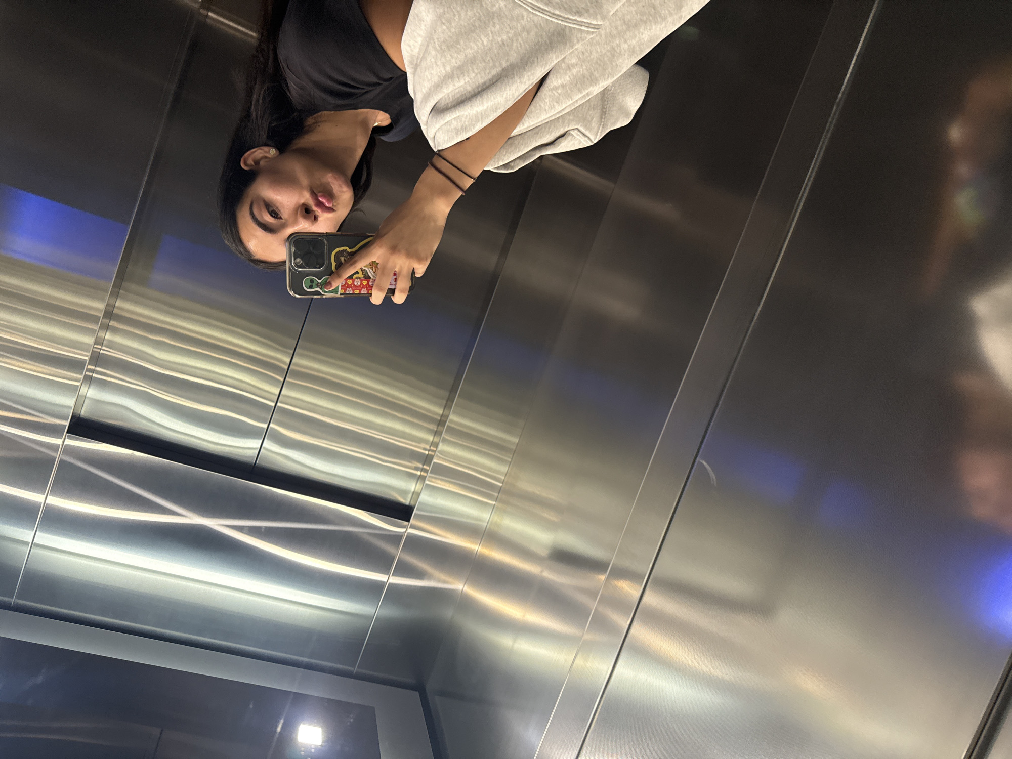 The image displays a selfie taken by a young woman reflected in the polished metal interior of an elevator. The photograph is oriented 90 degrees counter-clockwise, showing the woman from the chest up, appearing sideways. She has dark hair and is wearing a black t-shirt under a light grey hoodie. On her right wrist, two thin, dark bracelets are visible.
The woman holds a smartphone in her right hand, positioned to capture her reflection. The phone features a distinctive, colorful case adorned with numerous small, indistinct cartoon characters or stickers; no specific text is legible. Her lips are puckered in a selfie pose, looking towards the camera within the phone.
The elevator interior is characterized by brushed stainless steel walls that create a reflective surface. These walls show bright, elongated streaks and patterns from various light sources, including subtle blue and warm yellowish-white reflections, indicating a modern and well-lit environment. The reflection of the woman and her phone occupies the upper left portion of the frame, with the metallic surfaces and their light reflections filling the rest. This scene is located in Puebla, Mexico.