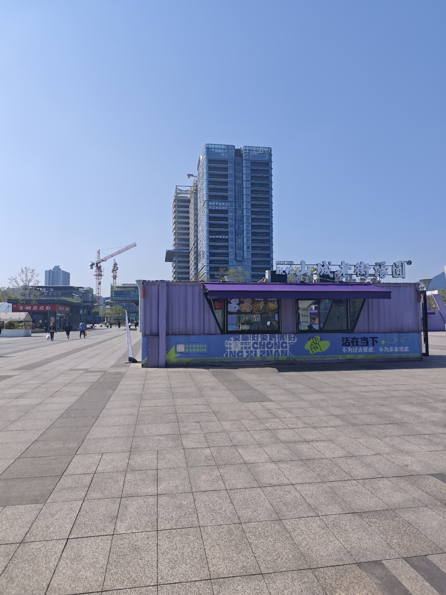 The image shows a wide paved plaza on a clear, sunny day in Shapingba District, China. In the foreground, a lavender-colored shipping container has been repurposed as a vendor stand. The stand's exterior is decorated with food pictures and Chinese characters. The text in English reads "CHONG ING XI ZHAN" is also on the container. Further text in Chinese suggests a food item (likely a dessert) is being sold, and a slogan about living in the present.
In the background, high-rise buildings are visible, including one under construction with two red and white cranes towering nearby. A few people are walking along the plaza. The ground is tiled with large, gray square pavers, and the sky is a clear blue.