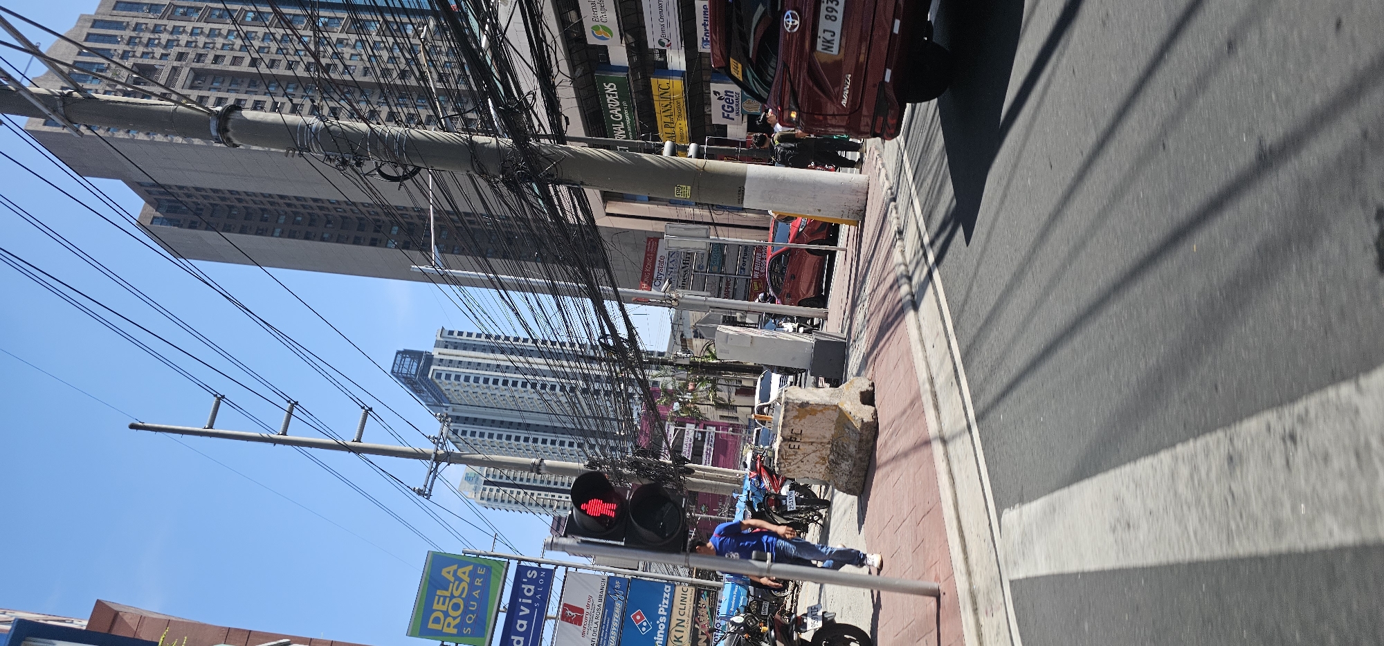 This is a street-level, wide shot of a busy intersection in Makati, Philippines. The scene is dominated by a dense network of overhead utility wires and several tall buildings, including one that appears to be a residential high-rise and another a commercial building with multiple floors. A pedestrian traffic light displays a red "don't walk" signal with a silhouette of a walking person. Several vehicles, including a red Toyota Avanza and other cars and motorcycles, are present on the road. On the sidewalk, there is a concrete barrier marked with "EPC", and a person in a blue shirt and jeans is walking. Several signs are visible, including "DELA ROSA SQUARE", "DAVID'S SALON", "MERCURY DRUG", "DOMINO'S PIZZA", and various other business advertisements and street signs. The sky is clear and blue, indicating a sunny day. The overall impression is one of urban density and activity.