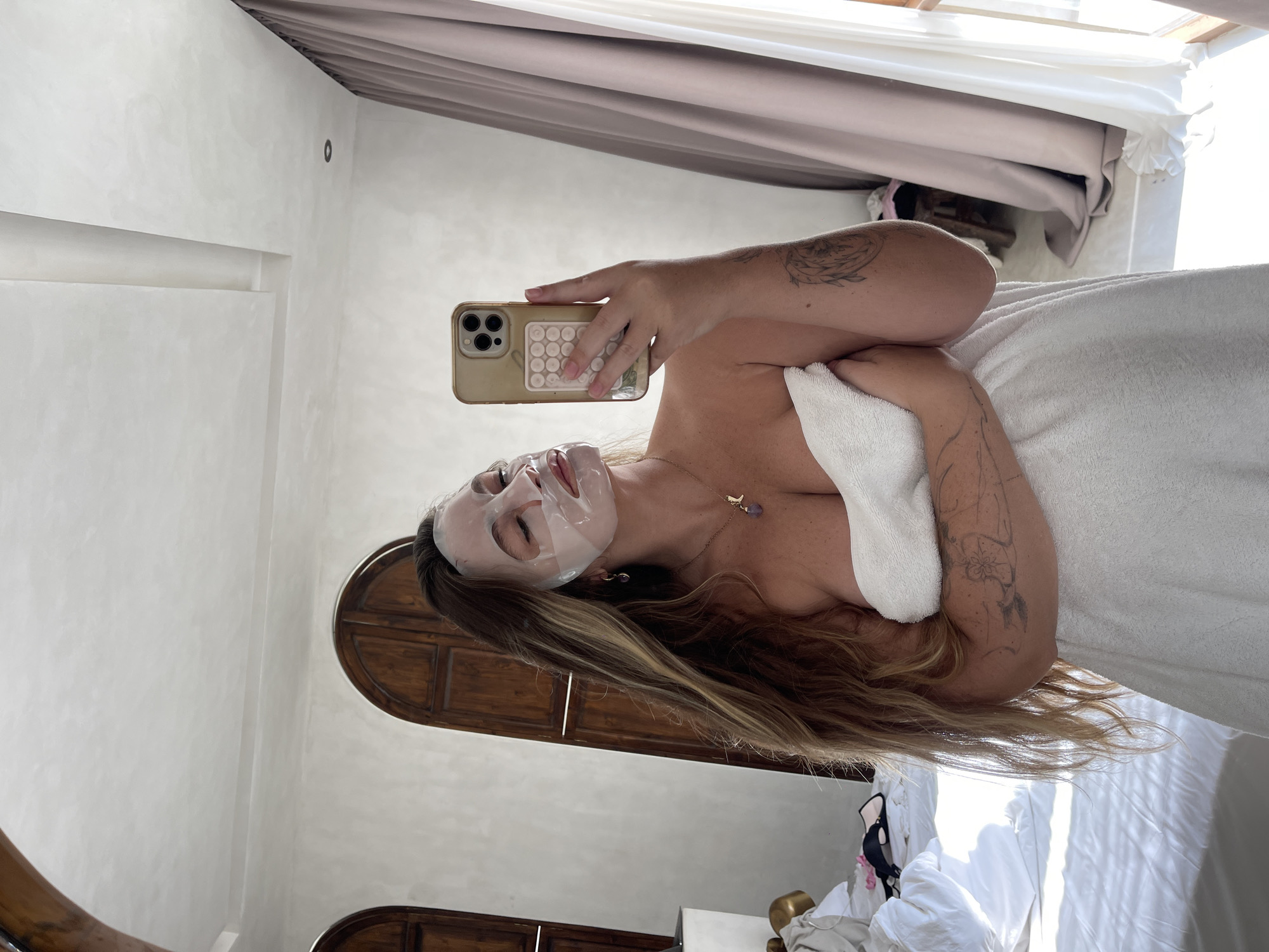 In a bedroom, a woman with long, blonde hair wears a white towel and a face mask. She holds a cell phone in her right hand, taking a selfie in a mirror. The room features a large bed with white linens on the left, and arched wooden doors and draped curtains on the right. Sunlight streams in from the right side of the frame.