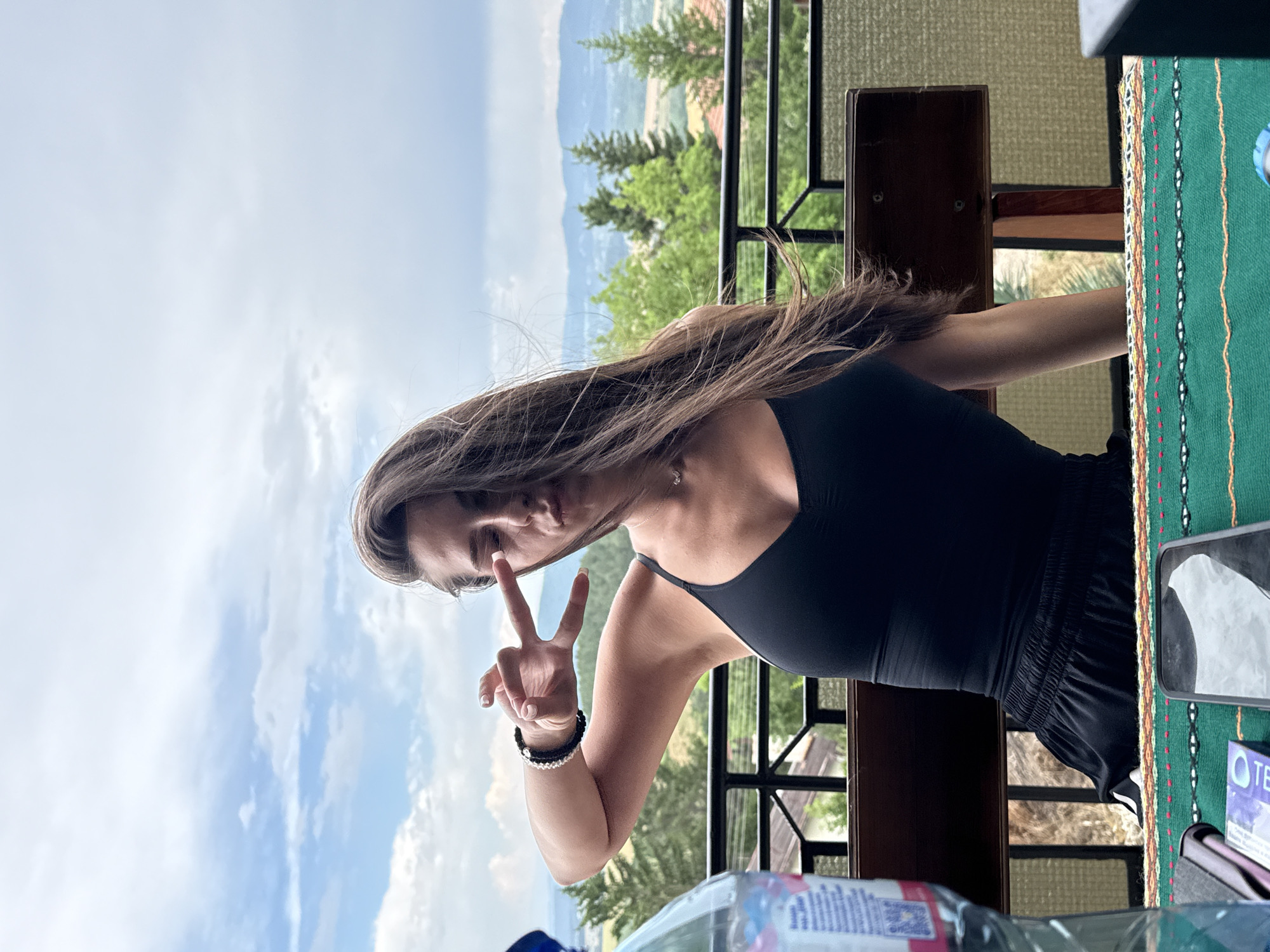 A young woman with long, brown hair is shown in a close-up shot, making a peace sign with her right hand. She is wearing a black tank top and black shorts. Her left hand is bent at the elbow, with her fingers extended. Her hair is blowing in the wind, obscuring part of her face. The background is a blurred landscape of green trees and mountains under a cloudy sky. The image appears to be taken outdoors during the daytime, with natural light. There is some text visible on a bottle in the foreground, but it is unreadable. A green, patterned textile is visible on the right side of the frame.