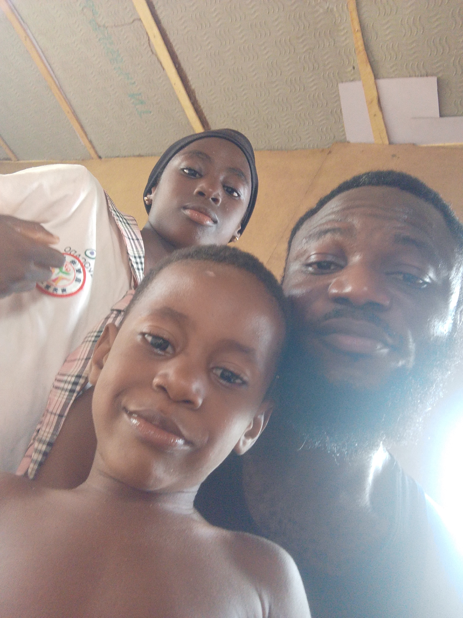 A close-up, low-angle selfie features three individuals of African descent. In the foreground, a young child, appearing to be bare-chested, looks towards the camera with a faint smile. Behind and to the left, a younger female wearing a white shirt, a plaid top, and a dark head covering also looks directly at the camera. She has small gold hoop earrings. On the right, an adult male with a beard and mustache is visible, his right side partially obscured by a strong glare from a light source. All subjects appear to be posing for the photograph.
The background reveals an indoor or covered outdoor setting with a textured ceiling made of patterned panels, possibly acoustic tiles, and prominent wooden beams running across it. In the upper right, a white piece of paper or board is attached to one of the beams.
Notable details include a circular emblem on the young female's white shirt, displaying a colorful design that includes what appear to be Chinese characters or symbols, along with some barely decipherable English-like text or numbers, possibly "ODA" or "021" and "0." Faint letters, "WTL," are visible printed on the ceiling panels in the upper left. The bright light source on the lower right creates a significant lens flare and obscures parts of the adult male and the immediate background. The subjects' expressions are generally neutral or subtly smiling. This photo was taken in Lokoja, Nigeria.