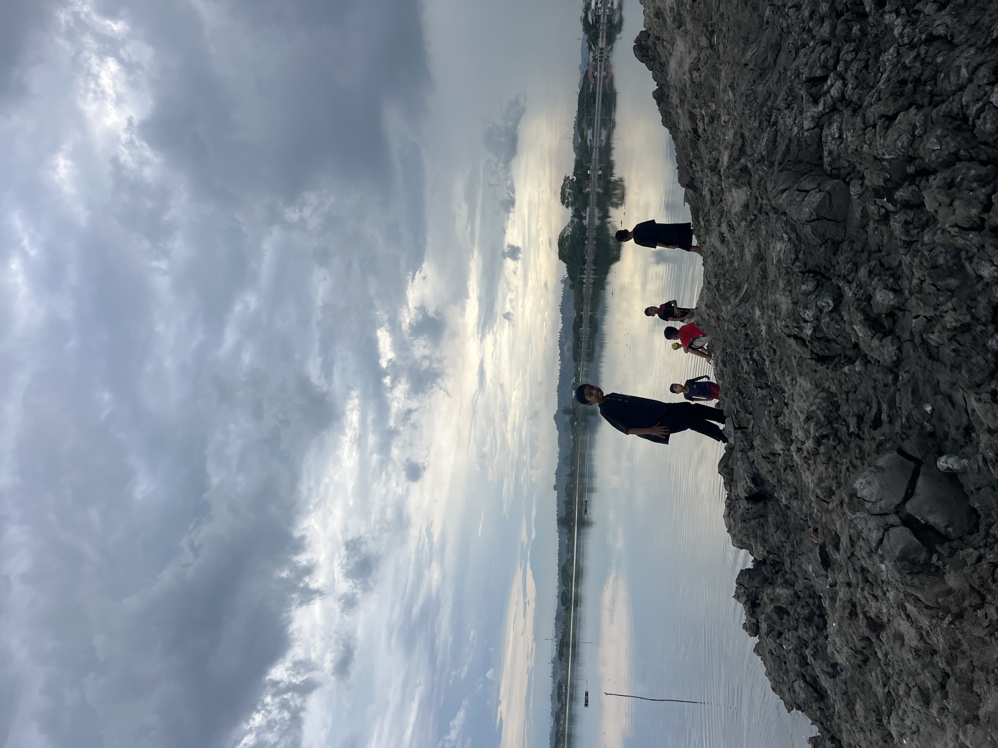 The image shows a group of people standing on a dark, cracked terrain that appears to be near a body of water, possibly a lake or a wide river, in Lhokseumawe, Indonesia. The sky is overcast with grey and white clouds, some of which are reflected in the water. The terrain has a rugged, uneven surface, suggesting dried mud or clay. A few people are clearly visible, one of them is wearing dark-colored clothing and stands more prominently in the foreground. A few other figures are standing further back, slightly obscured. The water is calm, and the opposite bank of the water body seems to have some vegetation and possibly some buildings.