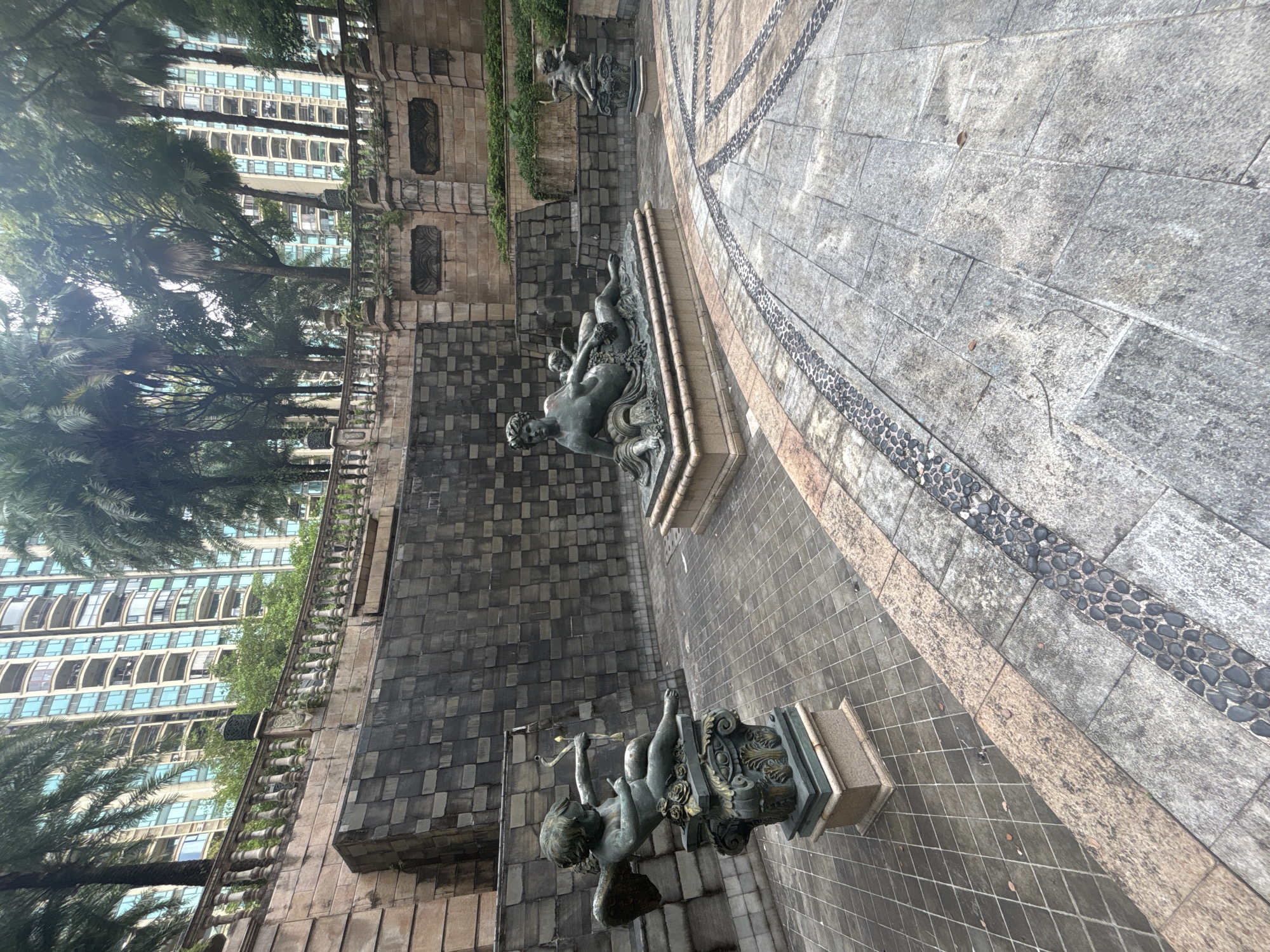 The image depicts a scenic courtyard in Nanhai District, China, characterized by classical statues and a tiered stone wall. In the foreground, a bronze statue of Cupid, complete with wings and a bow, rests on a decorative pedestal adorned with roses. To the right and further back, a larger bronze sculpture features reclining figures, possibly mythological deities, accompanied by cherubic forms. The courtyard floor is paved with interlocking stone tiles, featuring a decorative border of darker pebbles. The background reveals a dramatic stepped stone wall, partially overgrown with lush greenery. Behind the wall, towering apartment buildings and the fronds of palm trees suggest an urban setting. The overall atmosphere is one of tranquility and artistic appreciation, with the natural elements blending harmoniously with the man-made structures and sculptures.