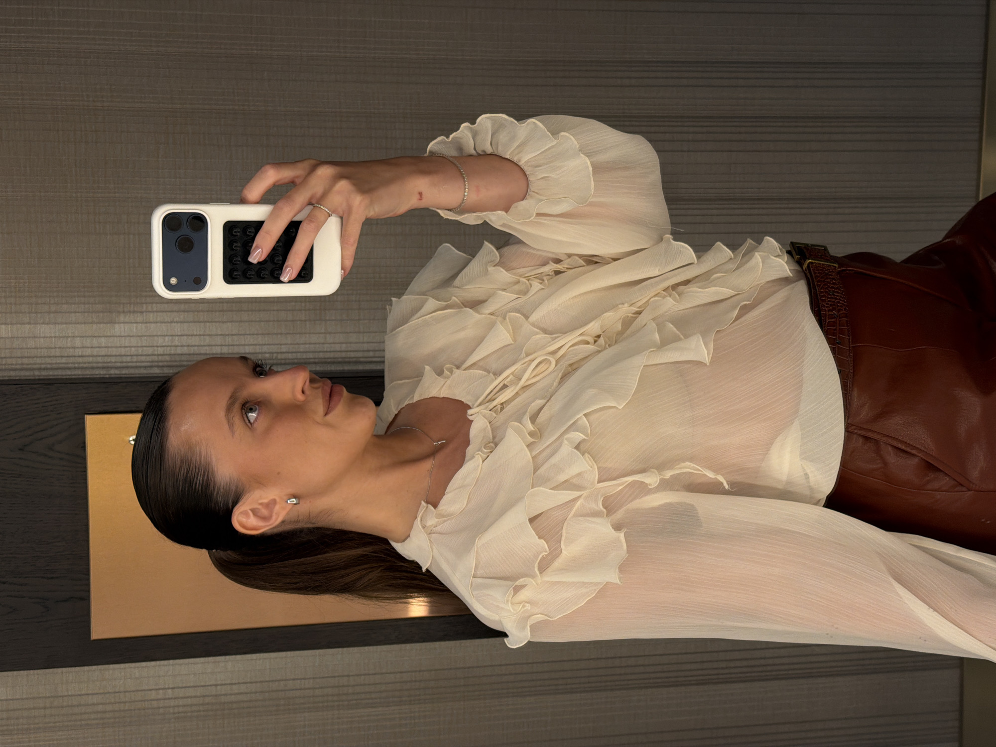 A woman with dark hair in a high ponytail takes a mirror selfie in what appears to be a hotel room in Budva, Montenegro. She is wearing a sheer, cream-colored blouse with ruffled detailing and a high neckline, paired with brown, faux-crocodile textured pants. A thin silver necklace with a small pendant is visible. Her right hand holds a white smartphone with a black pop socket and camera cover. Her left hand rests on her hip. The background consists of a dark, vertical-striped wallpaper and a framed rectangular object with a metallic copper finish.