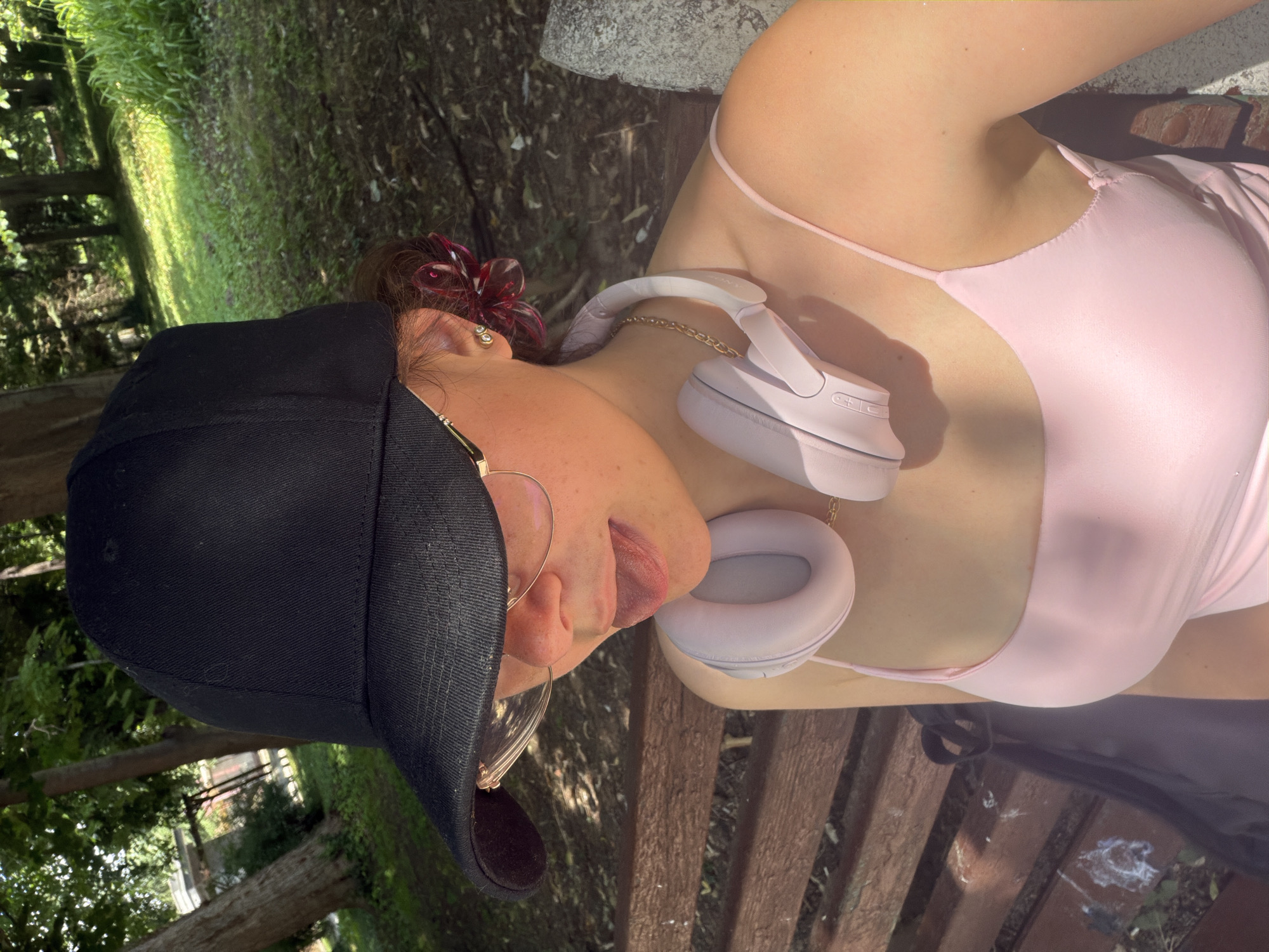 The image captures a partial view of an individual in an outdoor park setting in Pazardzhik, Bulgaria. A female subject is seen from the chest up, seated on a brown wooden park bench. She wears a black baseball cap pulled low over her face, obscuring her eyes. Gold-framed, clear-lensed glasses rest on her nose. Her tongue is slightly extended. Dark brown hair is visible behind her right ear, adorned with a reddish/burgundy, translucent, flower-shaped hair clip. A pair of light pink over-ear headphones, connected by a slender gold chain, are draped around her neck. She is wearing a light pink, thin-strapped top. The background features lush green trees and foliage under dappled sunlight. Green grass and exposed soil are visible on the ground. A portion of the wooden bench to the left of the subject shows white splatters, consistent with bird droppings. A dark bag rests on the bench next to her. A stone or concrete structure is partially visible on the far right.