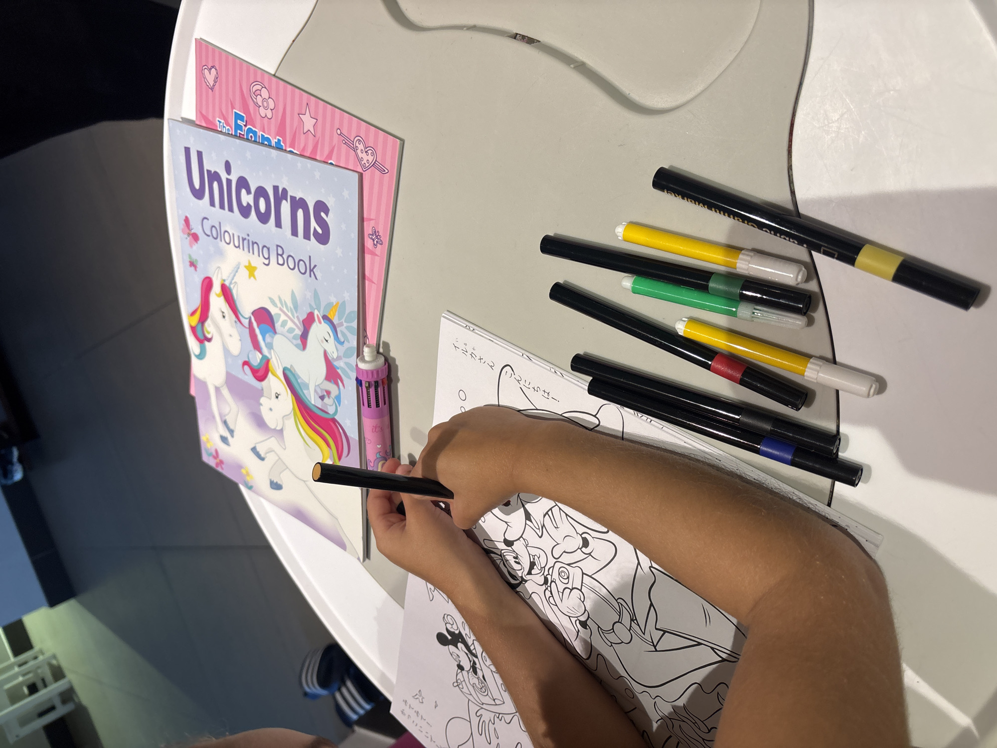 A child's hands are shown coloring a cartoon image in a coloring book. The child is holding a black marker. On the table are several other markers in various colors, a unicorn coloring book, and another pink coloring book with unicorn themed stickers. The scene is indoors, likely at a table, and the lighting suggests it is daytime. The environment does not provide specific clues to pinpoint the exact location within Dubai, United Arab Emirates.