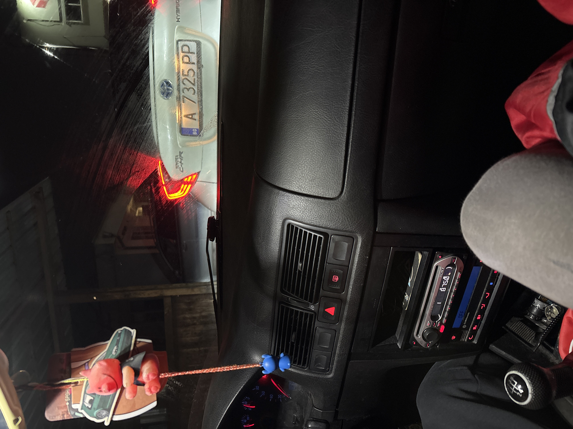 The image depicts the interior of a car, showing a dashboard with a radio and climate control panel. Hanging from the central air vent is an air freshener with blue, round objects on its ends and various small decorations attached. Through the windshield, a white Toyota C-HR Hybrid is visible, with the license plate "A 7325 PP" and the Bulgarian flag on the plate, indicating that the car and location are in Bulgaria. Based on the license plate code and other details, the location is likely in or around the city of Burgas, Bulgaria. A person's leg, covered in black fabric, is visible in the lower-right corner of the image.