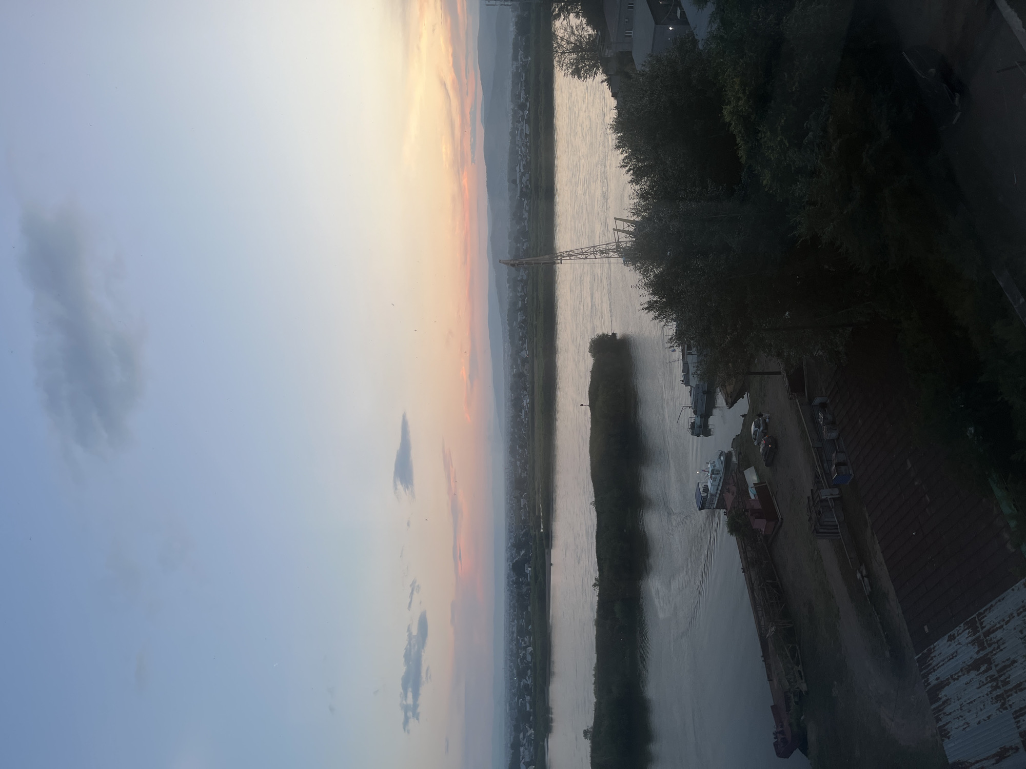 This is an aerial, high-angle, full shot of a waterfront scene at dusk. The sky transitions from a pale blue at the top to soft hues of pink and orange near the horizon, suggesting the sun has recently set. A wide, calm river occupies the mid-ground, flowing from left to right. On the opposite bank of the river, a town with houses and buildings is visible, nestled against a range of distant, hazy mountains. A large industrial crane stands near the right bank of the river. In the foreground, a dock area is visible with several boats, including a white passenger boat and a long barge. Two cars and a few people are on the dock. The foreground also shows rooftops of several low-rise buildings, some of which appear to be made of corrugated metal. The overall impression is a quiet, industrial port town at the close of day.