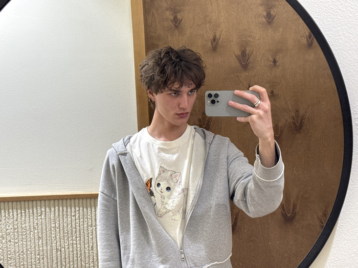 The image shows a young male individual capturing his reflection in a circular mirror for a selfie. He has medium-length, curly brown hair and blue eyes, and maintains a serious expression. He is dressed in a light-colored t-shirt featuring a graphic of a white kitten and a small orange butterfly, worn under an open, light gray zip-up hoodie with white trim on the cuffs and hem. His right hand holds a silver-colored smartphone, likely an iPhone due to the visible triple camera system, pointed towards the mirror. A silver-colored ring is visible on the ring finger of his right hand.
The environment comprises various wall finishes. To the left of the subject, a plain white wall is visible. Below it, a beige-toned textured wall with vertical fluting is present, separated by a light wood horizontal trim. The mirror's reflection reveals a section of a reddish-brown wood-paneled wall with an intricate, repeated, dark, abstract pattern resembling stylized eyes or feathers, positioned behind where the photo is being taken. This interior setting suggests a modern retail space, such as a clothing boutique or a changing room. The precise location within Tel-Aviv, Israel, cannot be determined from the visual information provided in the image.