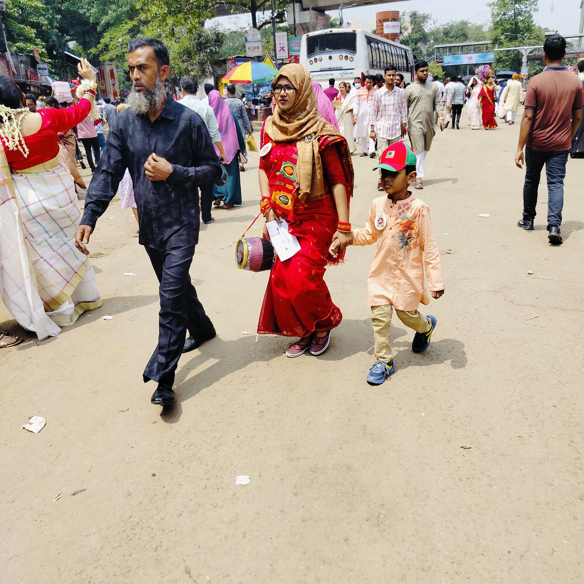 The image depicts a family of three walking amidst a crowd on a dusty street. The man, with a grey beard, wears a dark patterned shirt and dark pants. The woman, in a red sari and a brown headscarf, holds a small drum and some papers. A young boy in a peach-colored outfit and a red and green cap walks beside her, holding her hand.
The surrounding crowd includes individuals in various traditional and casual attire. A bus and several buildings are visible in the background, with signs in Bengali script. The location is Dhaka, Bangladesh, suggested by the language on the signs and the attire of the people. The surface of the street is sandy and covered in small pieces of litter.