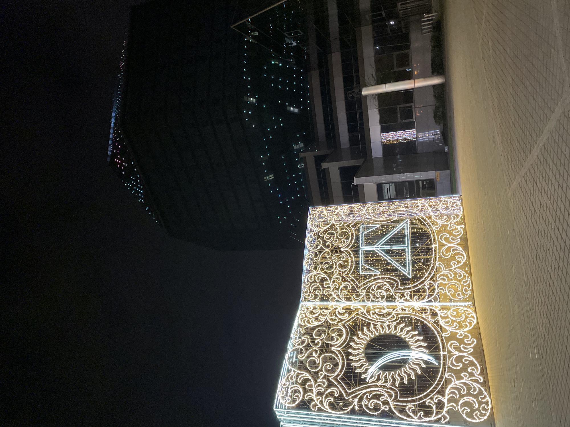 A nighttime cityscape in Minsk, Belarus, captures a vibrant urban scene dominated by illuminated buildings. In the foreground, the facade of a building is brilliantly adorned with elaborate decorative lights. These warm-toned string lights form intricate, swirling patterns that resemble vine-like scrollwork. Two prominent symbols are visible within these lights: an abstract, geometric design at the top, which could be interpreted as a stylized letter 'K' or a crown, and a circular motif below it, evocative of a sun or a celestial body with radiating rays and an arc. To the right of these bright decorations, the building's light-colored, textured facade, possibly made of bricks or panels, is subtly illuminated.
In the background and to the left, a tall, dark, modern skyscraper rises, its reflective surface sparkling with numerous small, scattered lights of various colors, possibly decorative elements or reflections from its windows, along with visible internal lights. Further to the right in the mid-ground, another contemporary building features dark, reflective glass windows that reveal interior lighting and structural details. The scene is set against a clear, dark night sky, indicating good weather. The extensive and intricate lighting suggests a festive occasion, likely during a holiday season. No people are visible in the image.