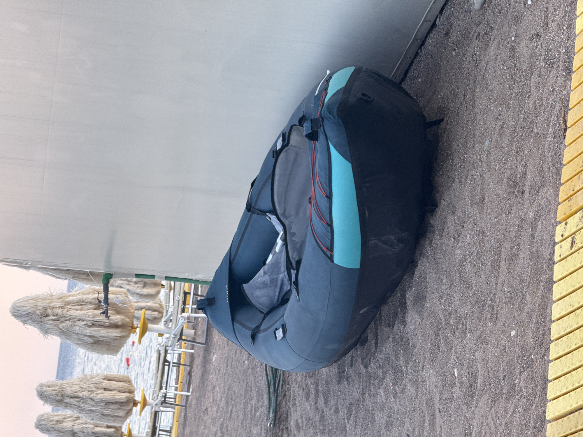 An inflatable dark blue and teal kayak rests on a sandy beach in Boreti, Montenegro. The kayak is positioned diagonally, with its bow pointing towards the right side of the frame, leaning against a white, corrugated wall. The beach is composed of small grey pebbles and sand. In the background, several large, straw-like umbrellas are set up, with lounge chairs visible behind them. The sea can be seen beyond the umbrellas, with calm water under a pale sky. A green hose lies on the sand to the left of the kayak. Yellow, slatted boardwalk planks are visible in the foreground.