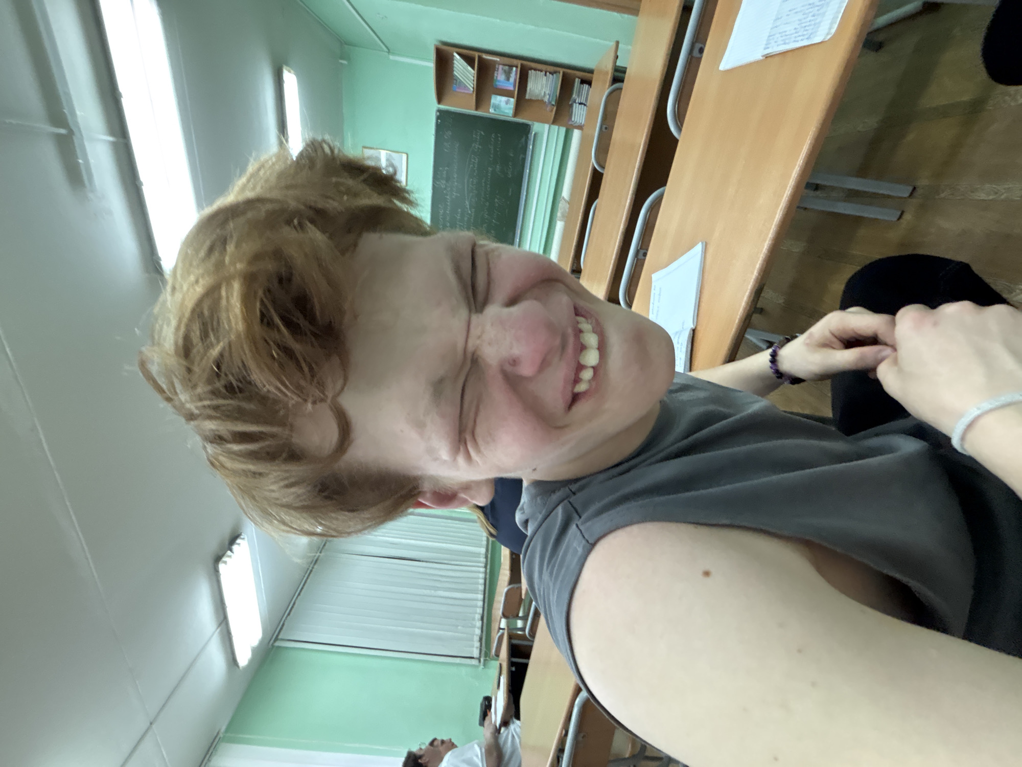 The image captures a playful selfie taken within what appears to be a classroom or lecture hall, oriented at a diagonal angle.
**Subjects:** The primary subject is a young person, likely a teenager or young adult, with reddish-brown, dishevelled hair. They are positioned in the foreground, looking directly at the camera with a wide, squinting smile that conveys joy or amusement. Their eyes are almost closed due to the intensity of their expression. They are wearing a dark grey sleeveless top, and on their left wrist, a purple beaded bracelet is visible, along with a lighter-colored bracelet on their right wrist. In the background, partially visible towards the bottom left, is another person sitting at a desk, wearing a white shirt and looking upwards, though their face is not clearly discernible.
**Scene and Setting:** The setting is an indoor classroom environment. Light brown wooden desks with metal frames are arranged in rows, with some papers scattered on them. The walls are painted a light green color. On the back wall, a blackboard with handwritten text, appearing to be in Cyrillic script, is visible. Above the blackboard, shelves hold books or binders. Fluorescent strip lights illuminate the ceiling. A window covered with white blinds is also present.
**Activity or Event:** The main activity is the taking of a selfie, characterized by the subject's expressive and joyful pose. The presence of multiple desks, a blackboard, and books suggests a learning or academic context, possibly during a break or after a class.
**Notable Details:** The strong fluorescent lighting indicates an indoor setting. The clear, bright light doesn't strongly suggest a specific time of day, but it is likely daytime. The Cyrillic script on the blackboard is a significant location cue, suggesting the setting is in a country where Cyrillic is used (e.g., Russia, Ukraine, Belarus, etc.). The overall mood is lighthearted and cheerful due to the subject's expression.
**Visible Text:**
* On the blackboard: Handwritten text in Cyrillic script.
* On papers on desks: Handwritten text is visible but illegible.