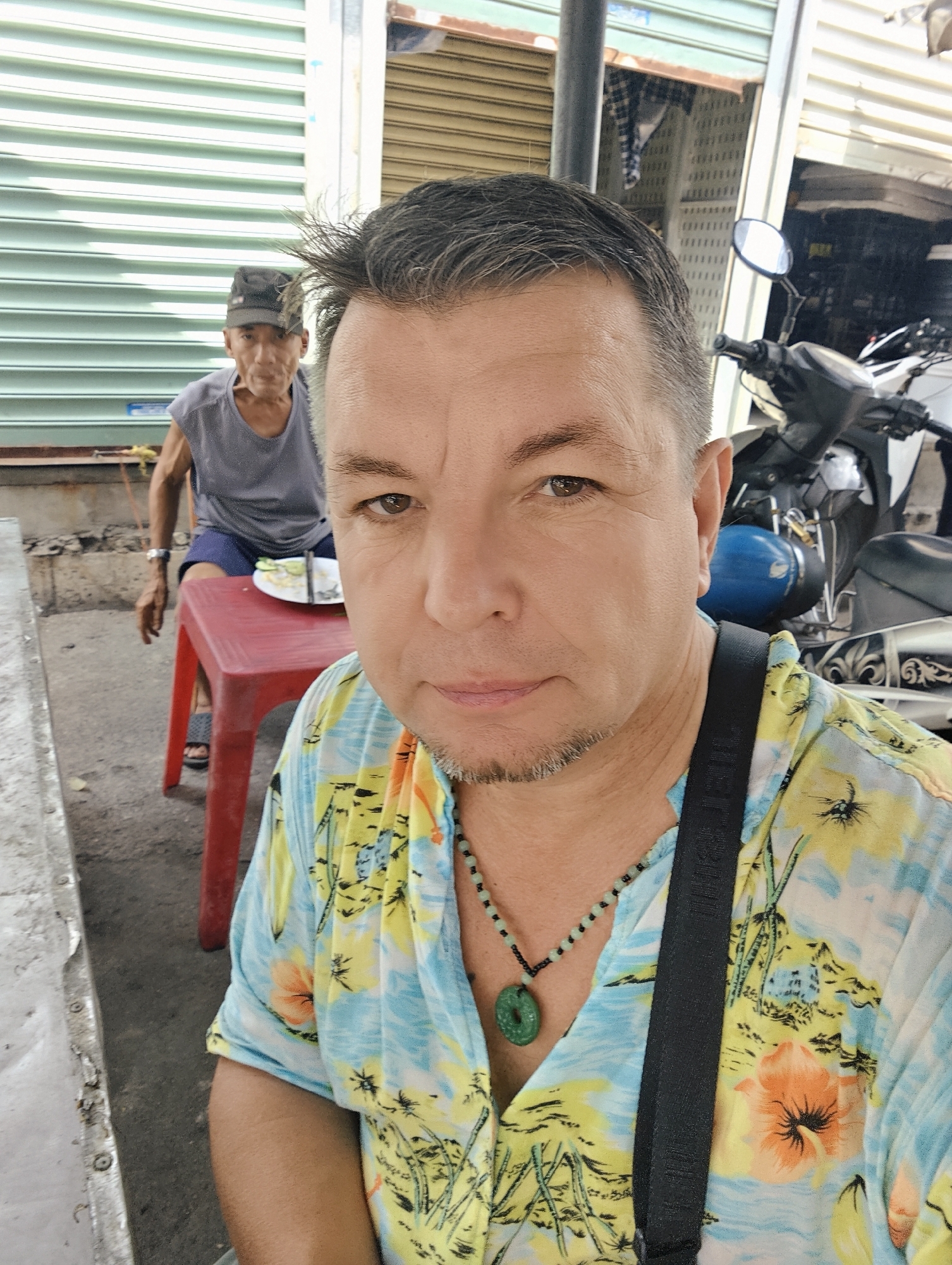 In Nha Trang Ward, Vietnam, two individuals are present in an outdoor setting, possibly a street-side establishment. The foreground is occupied by a man taking a selfie, his upper body and face dominating the frame. He is wearing a brightly colored floral shirt and a necklace with a green pendant. To the left of the frame, a metal surface is visible. In the background, an older man sits on a red plastic stool, wearing a dark baseball cap and a gray t-shirt. He is seated at a small table with a plate of food. Behind him, a corrugated metal wall and a parked motorcycle with a blue helmet are visible. The overall environment suggests a casual, everyday scene in a Vietnamese urban area.