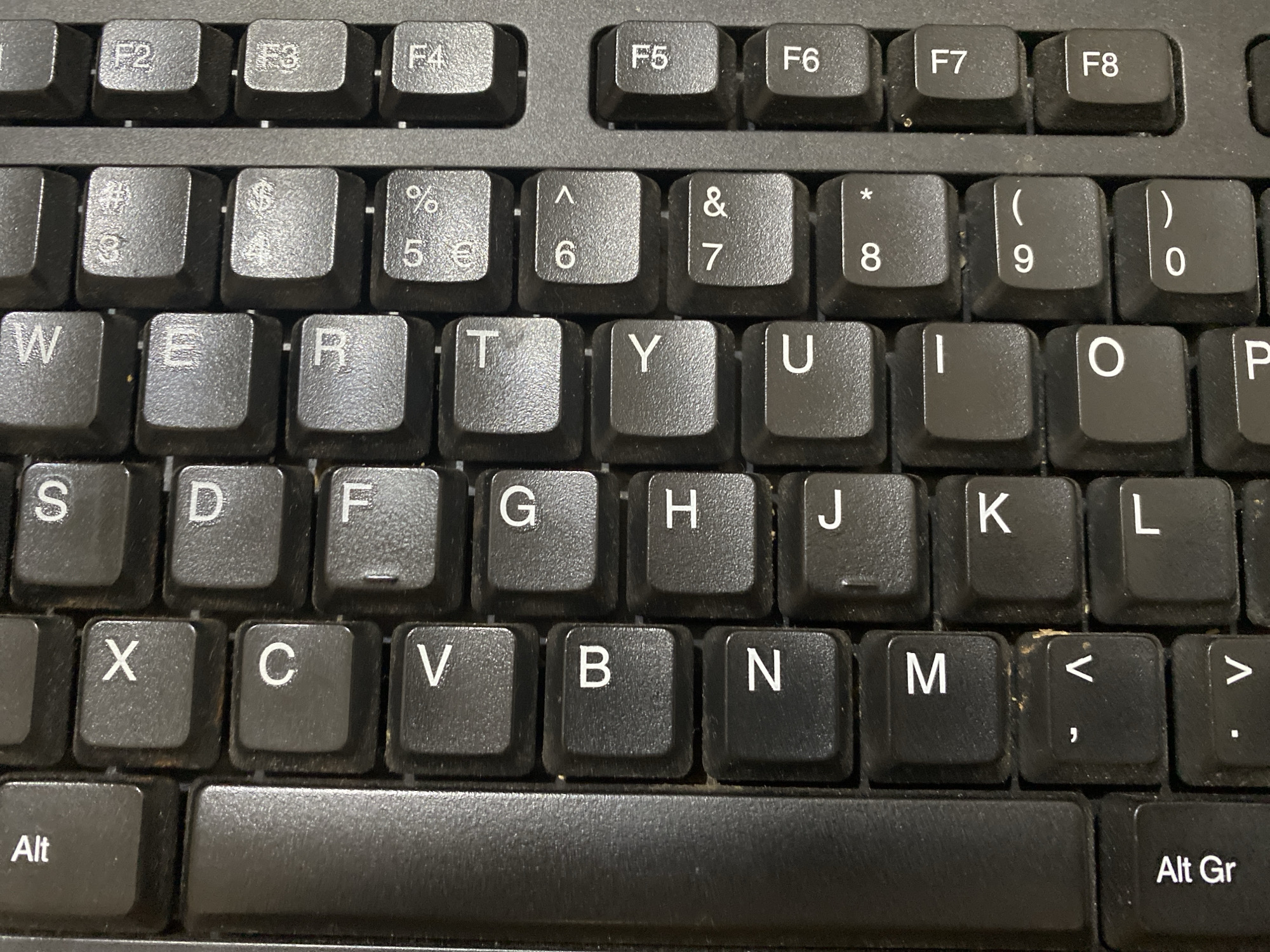 The image shows a close-up, high-angle view of a black computer keyboard. The keyboard appears to be well-used, with visible dust and debris between the keys, particularly in the crevices. The keys themselves are mostly clean, displaying standard QWERTY layout with letters, numbers, and function keys (F1-F8) clearly visible. The image focuses on the upper and middle rows of keys, including the number row with symbols above them, the top letter row (QWERTYUIOP), and the home row (ASDFGHJKL). The bottom row shows the 'Alt' key. The overall environment is a nondescript, indoor setting implied by the presence of the keyboard, but no other environmental details are discernible. There are no people, animals, or any contextual clues that would suggest a specific location like Dabasah, Indonesia. The image is solely a depiction of a keyboard.