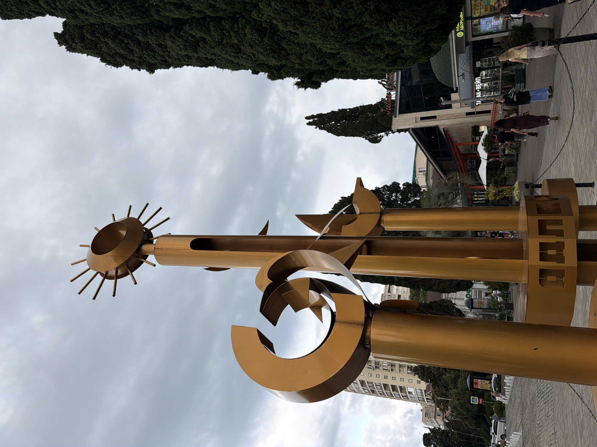 This media file captures an outdoor scene with a prominent golden sculpture in the foreground. The sculpture features a circular element with radiating spikes, resembling a sun, and other abstract metallic forms. The image is oriented vertically, with the sky occupying a significant portion of the frame. In the background, there are buildings, trees, and several people walking on a paved area. The weather appears overcast, with a cloudy sky. The lighting suggests it could be daytime, possibly late afternoon or early morning. Visible text on a building in the background includes "ТАК-ВЕЙЛ" and another sign with Cyrillic letters that may read "РИО". There are also some circular signs on poles. The overall setting appears to be an urban environment or a public space, possibly a promenade or a commercial area, given the presence of shops and pedestrians. The people are casually dressed and seem to be engaged in everyday activities like walking and shopping.