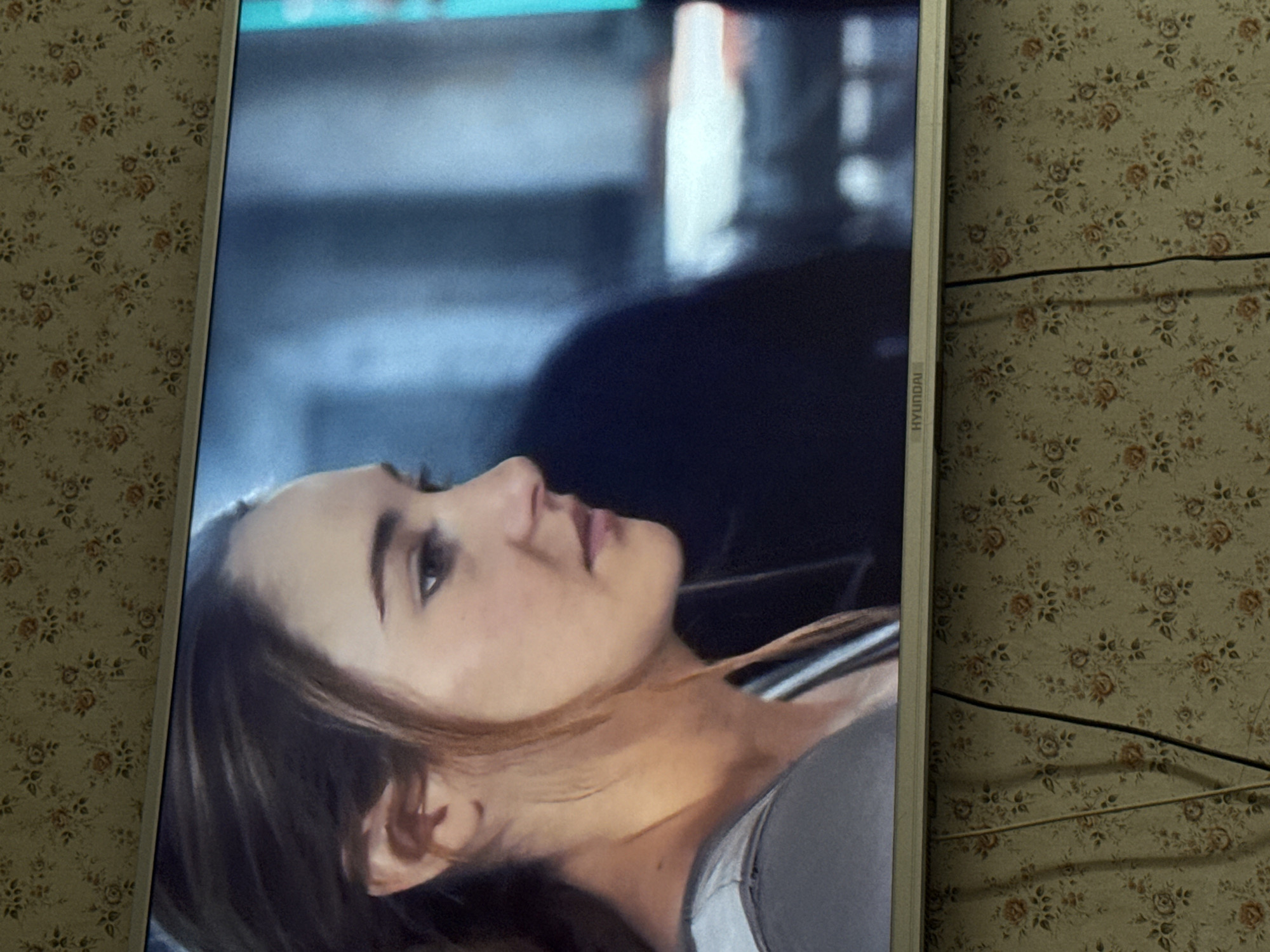 A television displaying a close-up of a woman's face is positioned against a wall covered in a patterned wallpaper. The woman has light skin, dark hair, and is looking upwards. The television is identified as a Hyundai based on visible branding. The image does not provide sufficient information to determine a specific location within the city of Mytishchi, Russia.