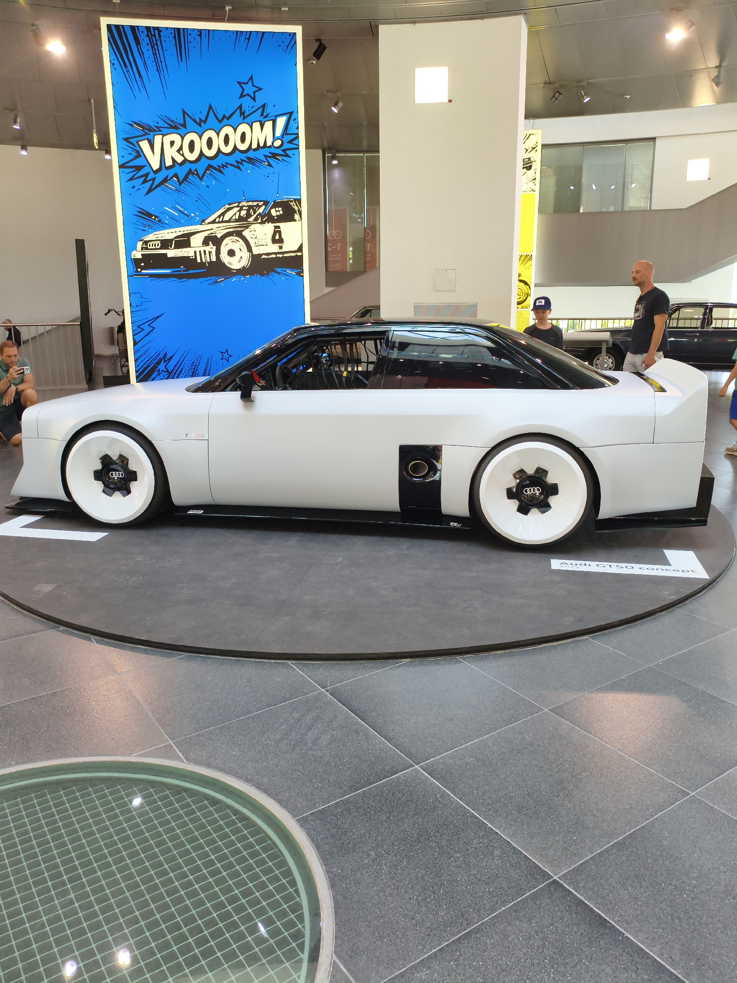 This image depicts the Audi GT50 concept car on display, likely within the Audi Forum in Ingolstadt, Germany, a common location for such exhibitions. The car, a striking white sports coupe with black accents, is positioned on a circular display platform with a dark grey tiled floor surrounding it. The platform features a white border and a small sign indicating "Audi GT50 concept".
Behind the car, a large, vertical graphic panel is prominently displayed. This panel features a blue comic-book-style background with black starbursts and an outline of a racing car with the word "VROOOOM!" in white lettering.
In the foreground, a section of a glass floor with a green grid pattern is visible. Several people are present in the scene. To the left, a person is crouching and taking a photo of the car with a smartphone. To the right, two men are standing and observing the exhibition. One man, wearing a dark t-shirt and light shorts, is facing the car. A young boy wearing a baseball cap stands near him, also looking towards the car. Further in the background, another car, appearing to be a classic black Audi, is partially visible. The environment suggests an indoor exhibition space with a high ceiling and diffused lighting, typical of a museum or automotive showroom.