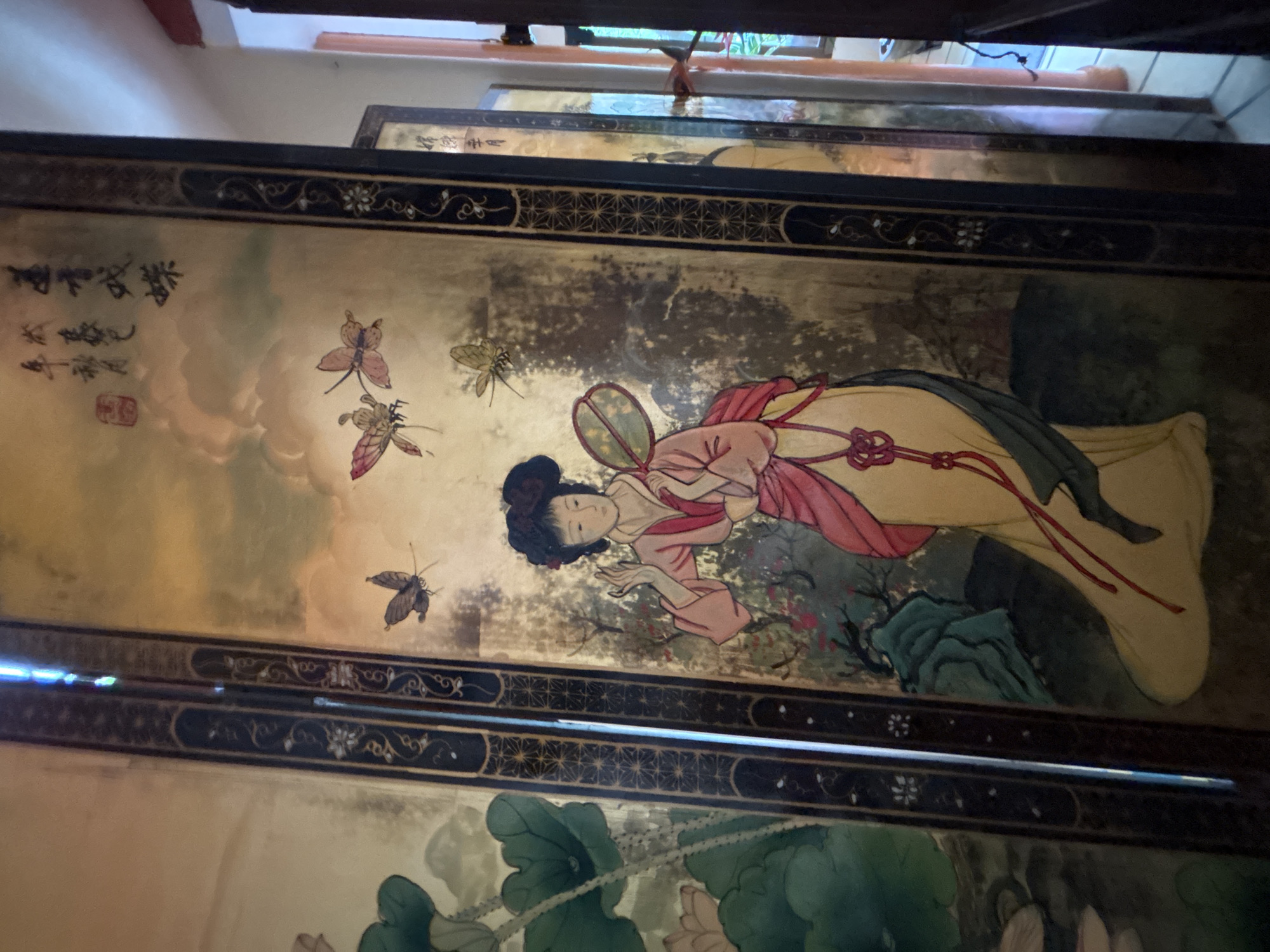 A folding screen, featuring a painted scene of a woman in traditional East Asian attire, is positioned in a room. The woman is depicted with dark hair styled in an updo, a pink outer robe over a yellow inner garment, and is holding a fan. Butterflies are painted in the background, along with foliage and clouds. The screen is bordered by decorative dark panels with inlaid patterns. In the background, other folding screens and a doorway can be seen. The lighting suggests an indoor environment, possibly a home or shop in Chalong, Thailand, with natural light entering from the side.