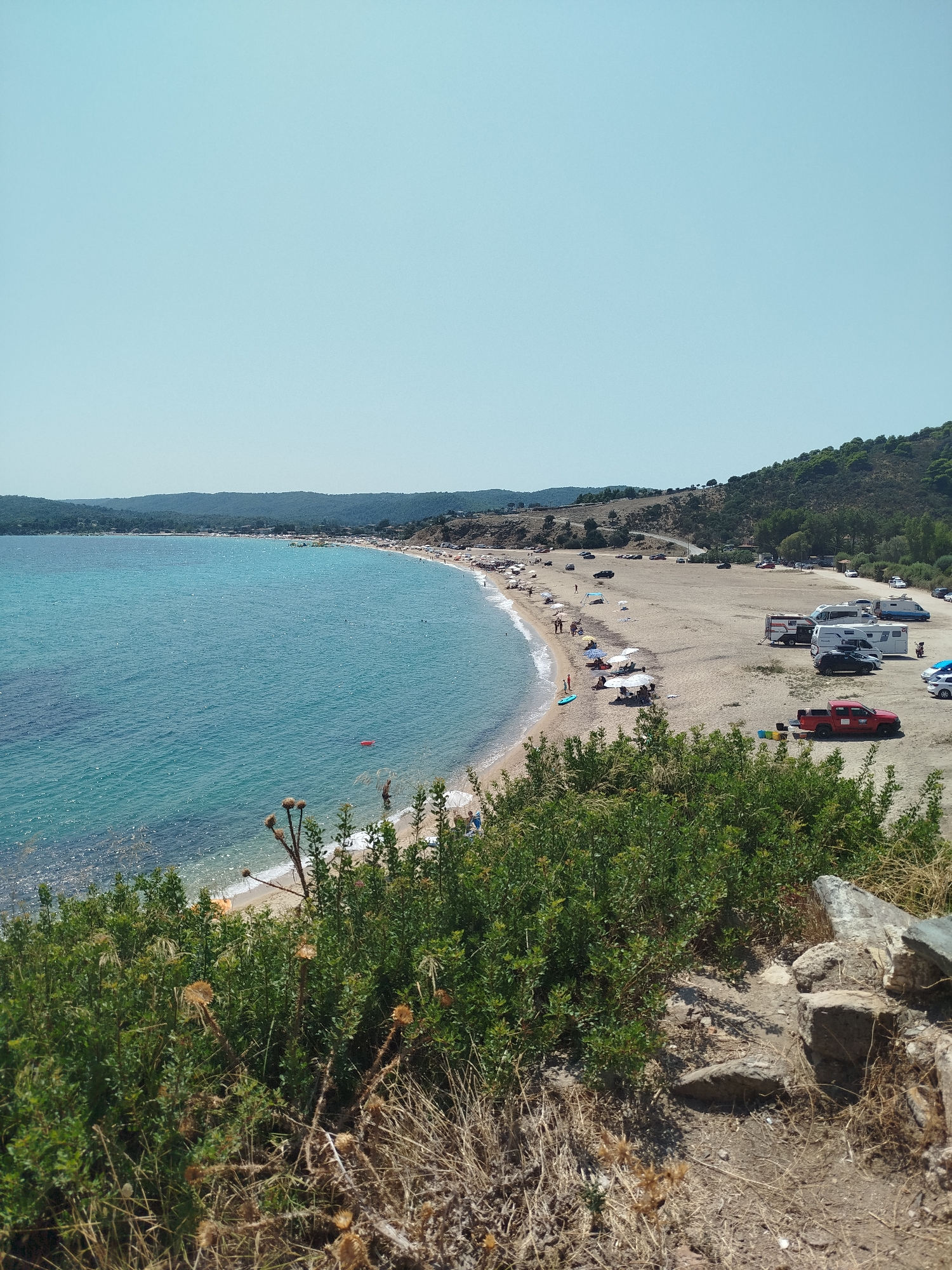 This is an aerial, wide-angle shot of a crescent-shaped sandy beach on the coast of Sithonia Municipal Unit, Greece. The beach is lined with green vegetation in the foreground and is dotted with several beach umbrellas, people, and vehicles. The turquoise water is calm and clear, with a few people swimming or wading near the shore. In the background, there are hills covered with green trees and a clear blue sky. A road winds along the top of the beach, and several RVs and cars are parked on the sand. The overall scene depicts a popular beach destination on a sunny day.