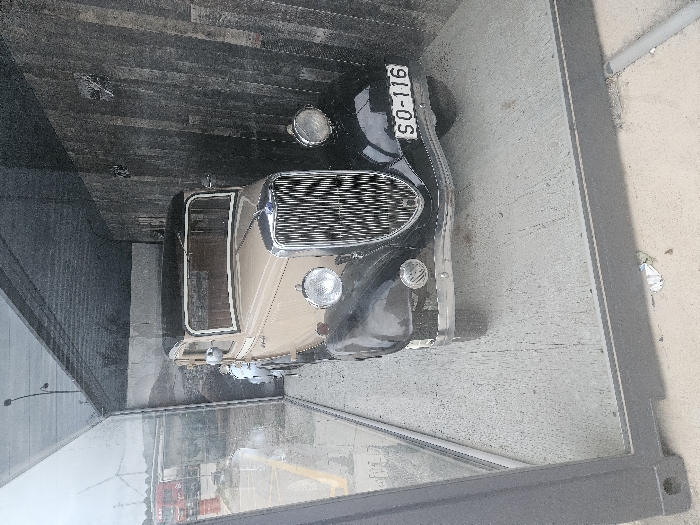 A vintage two-tone sedan, featuring a light tan body with black fenders, running boards, and roof, is displayed within a transparent glass and dark gray metal-framed enclosure. The vehicle exhibits a prominent vertical chrome grille, two large circular headlights, and a front-mounted license plate reading "SO-116". The car rests on a concrete slab inside the enclosure. The interior rear wall behind the car displays a large photographic mural depicting a rugged, hilly landscape. The roof and right interior wall of the enclosure appear to be constructed from dark, possibly corrugated metal or weathered wooden planks.
Through the left glass panel of the enclosure, an outdoor environment is visible under an overcast sky. This includes a paved area marked with yellow lines, a concrete bridge or overpass structure, and reddish-orange industrial containers or structures in the mid-ground. In the far distance, several large wind turbines are discernible. A street lamp extends from the upper left. Crumpled white litter is present on the ground outside the enclosure's base. No individuals are depicted. This scene is located in the city of Tudela, Spain.