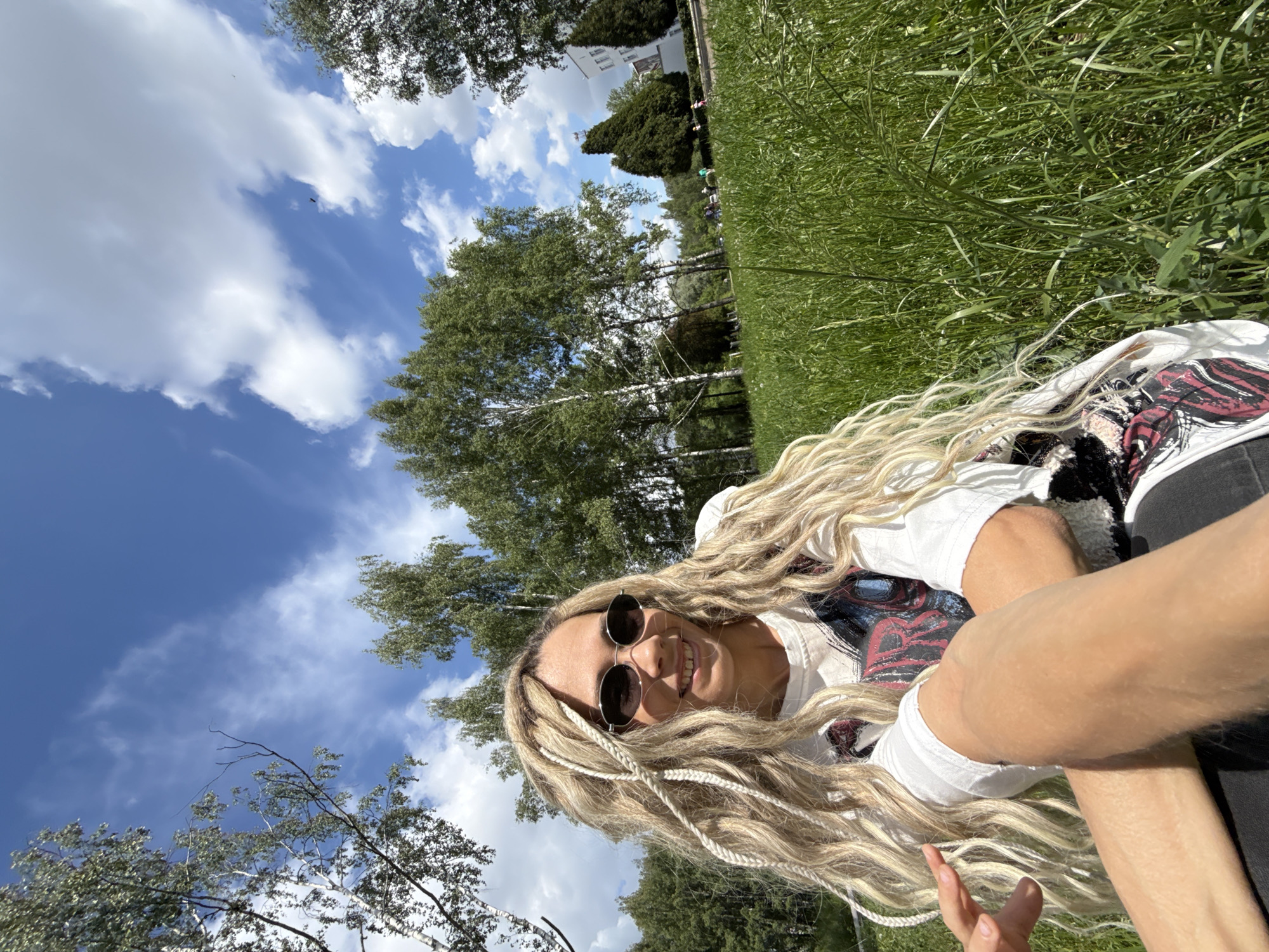 A young woman is captured in a selfie, lying on her back in tall, green grass, looking up at the camera with a joyful expression. She has long, blonde hair, some of which is styled in braids, and she is wearing round, dark sunglasses. Her smile is visible, indicating happiness and relaxation. She is dressed in a white t-shirt featuring a graphic design with red and black elements, possibly depicting a face or mask, and dark pants.
The setting is outdoors in a park or field in Rusanovo, Russia, under a bright daytime sky. The weather is clear and sunny, with scattered white, cumulus clouds against a vibrant blue background. Lush green grass fills the foreground, while the background features numerous tall trees, including birches with their distinctive white bark. Beyond the trees, indistinct figures of people and hints of buildings can be seen in the distance, suggesting a populated area adjacent to the natural environment. The image is taken from a low angle, emphasizing the sky and the woman's upward gaze.