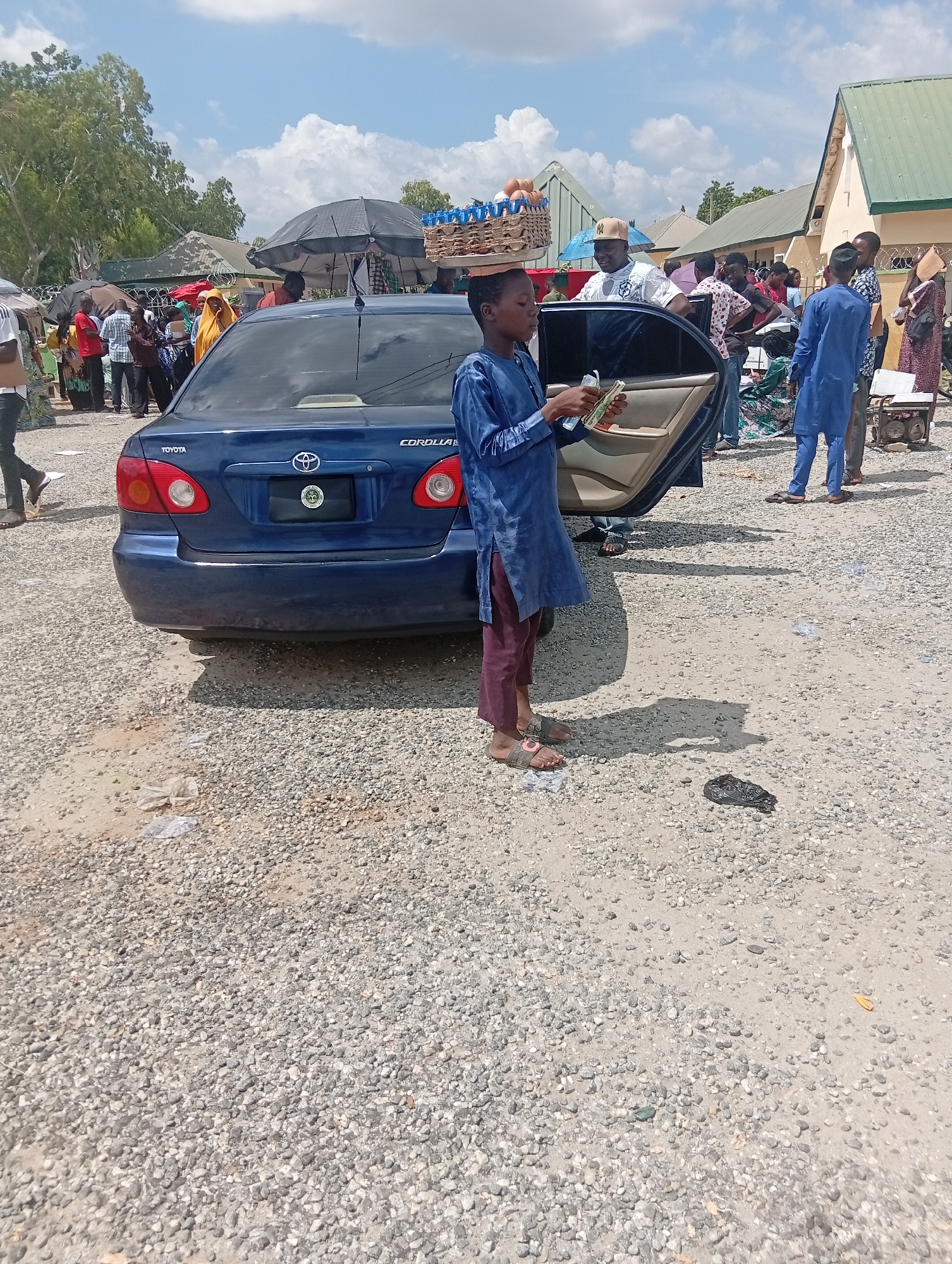 The image depicts an outdoor scene, possibly a market, with a blue Toyota Corolla parked on a gravel and dirt lot. A young boy, dressed in a blue tunic and dark pants, stands by the open driver's side door of the car, holding a wad of cash. He is looking towards the camera, and his feet are bare except for sandals. Behind him, a basket of eggs is precariously balanced on someone's head.
In the background, a crowd of people are gathered, some under umbrellas. Buildings with green and beige roofs are visible, along with trees and a cloudy sky. A generator is situated to the right of the car. The overall impression is of a busy, informal commercial area.
