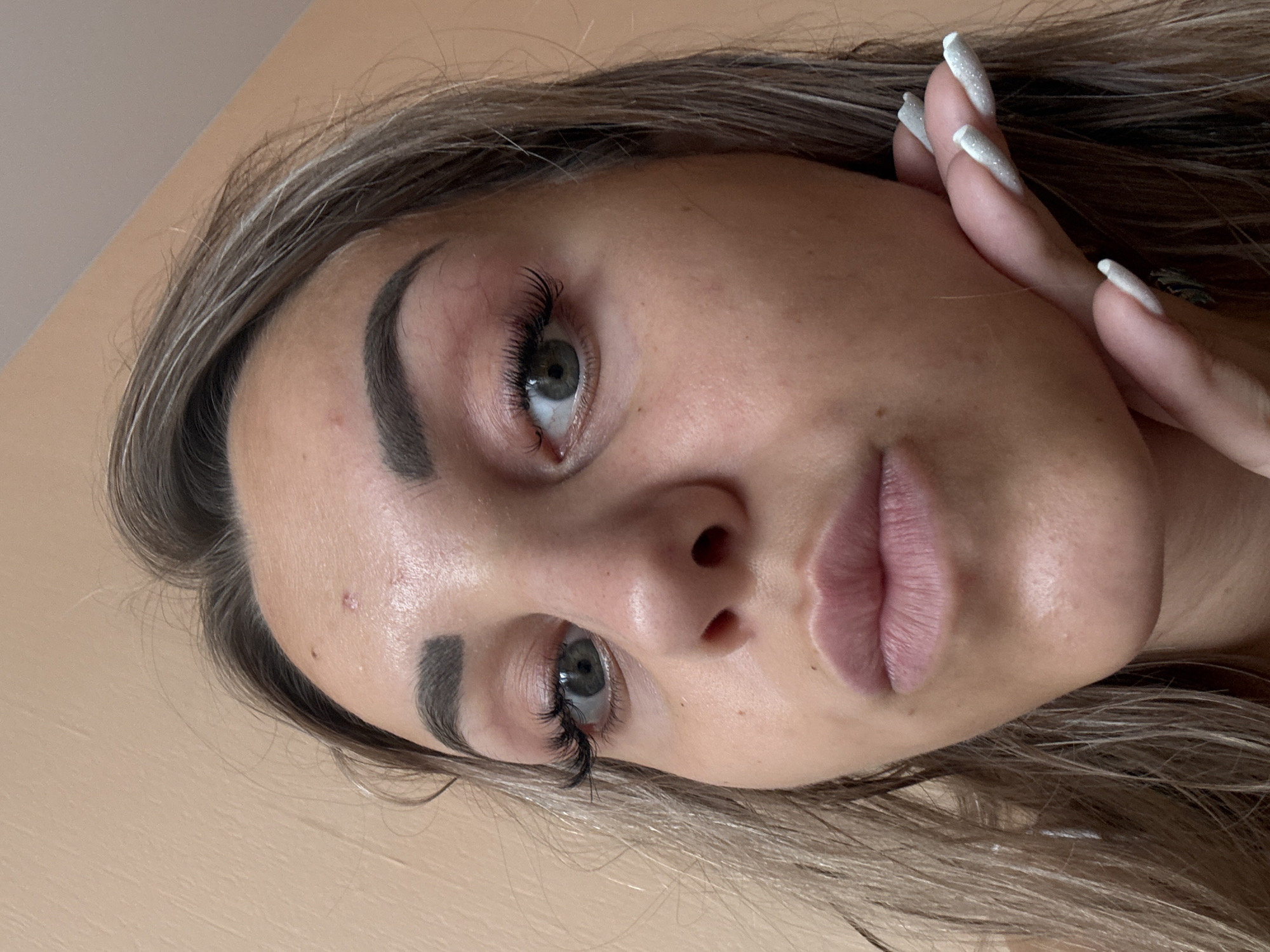 The image is a close-up, high-angle shot of a young woman's face. Her head is tilted, and her left hand is touching her chin. She has long, light brown hair with blonde highlights, dark eyebrows, and visible false eyelashes. Her eyes are green, and her lips are slightly parted. There appears to be a small blemish or redness on her forehead. The background is a plain, peachy-colored wall. The image does not contain any elements that would allow for identification of its location in Kholmsk, Russia, or any other specific city.