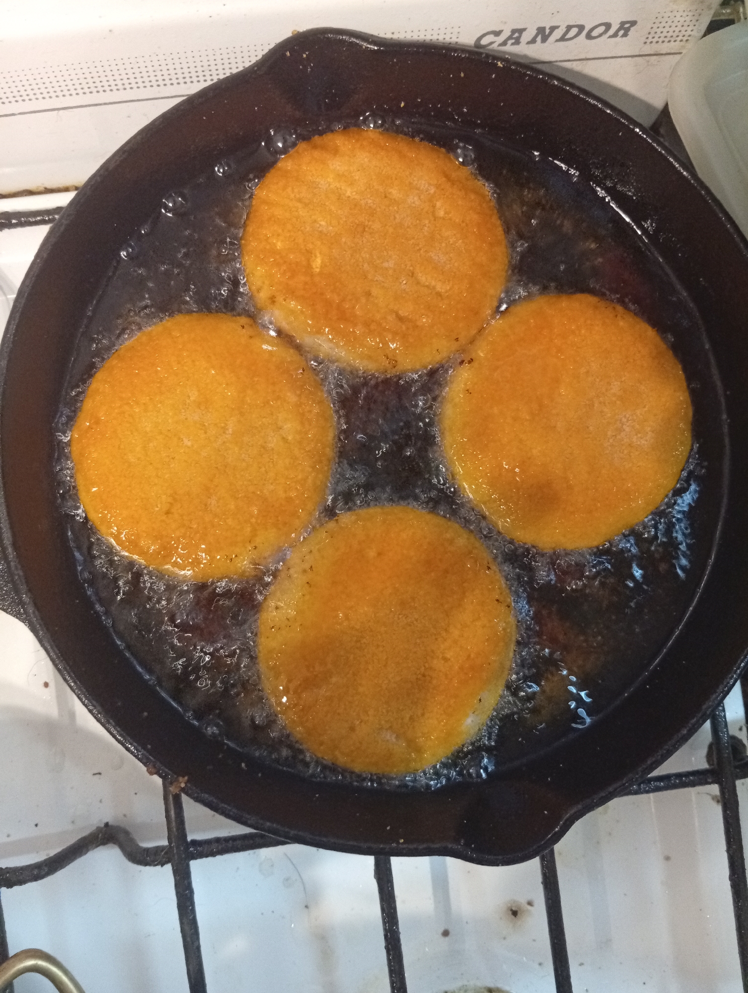 An overhead shot captures four circular, orange-gold colored patties or fritters actively frying in bubbling oil within a dark cast iron skillet. The skillet occupies the central and lower portions of the frame, positioned on a stovetop with black metal grates visible at the bottom and right edges. The food items are arranged in a roughly square pattern within the skillet, each surrounded by a vigorous simmer of hot oil.
In the background, behind the skillet, part of a white appliance, likely a stove or oven, is visible. On this appliance, the word "CANDOR" is clearly printed in dark, capital letters, suggesting a brand name. The surface of the appliance also features a textured pattern of small, raised dots.
The scene depicts a cooking activity, specifically deep-frying or pan-frying, taking place indoors. The bubbling oil indicates high heat and active cooking. The overall impression is one of homemade food preparation in a kitchen, consistent with everyday life in a place like Buenos Aires, Argentina, given "CANDOR" is a known Argentinian home appliance brand. The time of day and weather are not discernible.