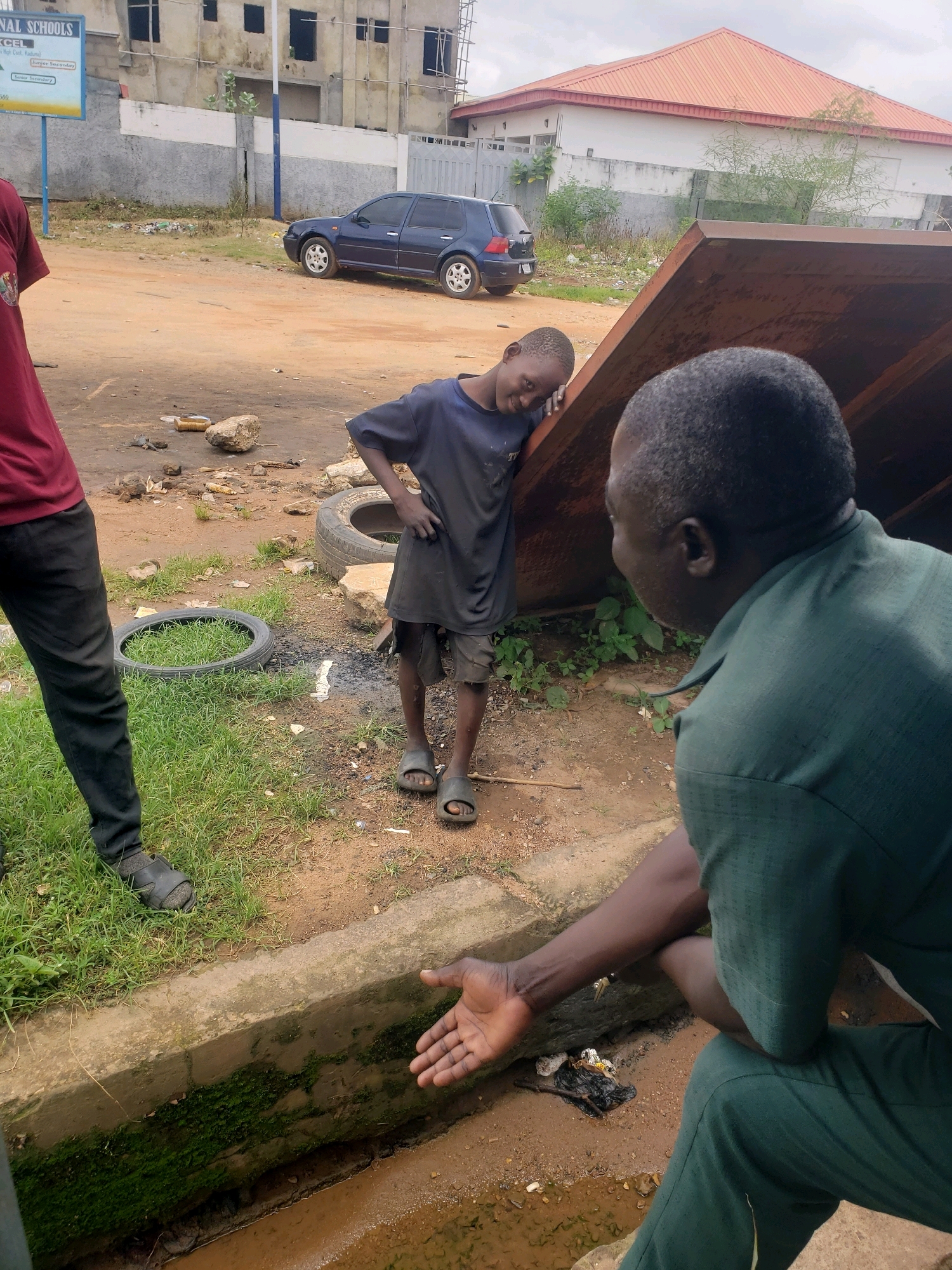 This image depicts a street scene in Sabon Tasha, Nigeria. In the foreground, a man in a green collared shirt and pants is kneeling, facing away from the camera and towards a young boy. His left hand is extended towards the boy, palm open. The boy, wearing a dark t-shirt and torn shorts with sandals, is leaning against a large, rusty metal object, looking towards the kneeling man with a slight smile. To the left of the boy, a dark-colored Volkswagen Golf is parked. Behind the car, a partially constructed building stands, with scaffolding visible. The ground is a mixture of dirt and grass, with discarded tires and debris scattered around. A concrete drainage channel runs along the bottom of the image, with moss growing on its sides and murky water flowing within. The overall impression is one of a utilitarian, somewhat unkempt urban environment.