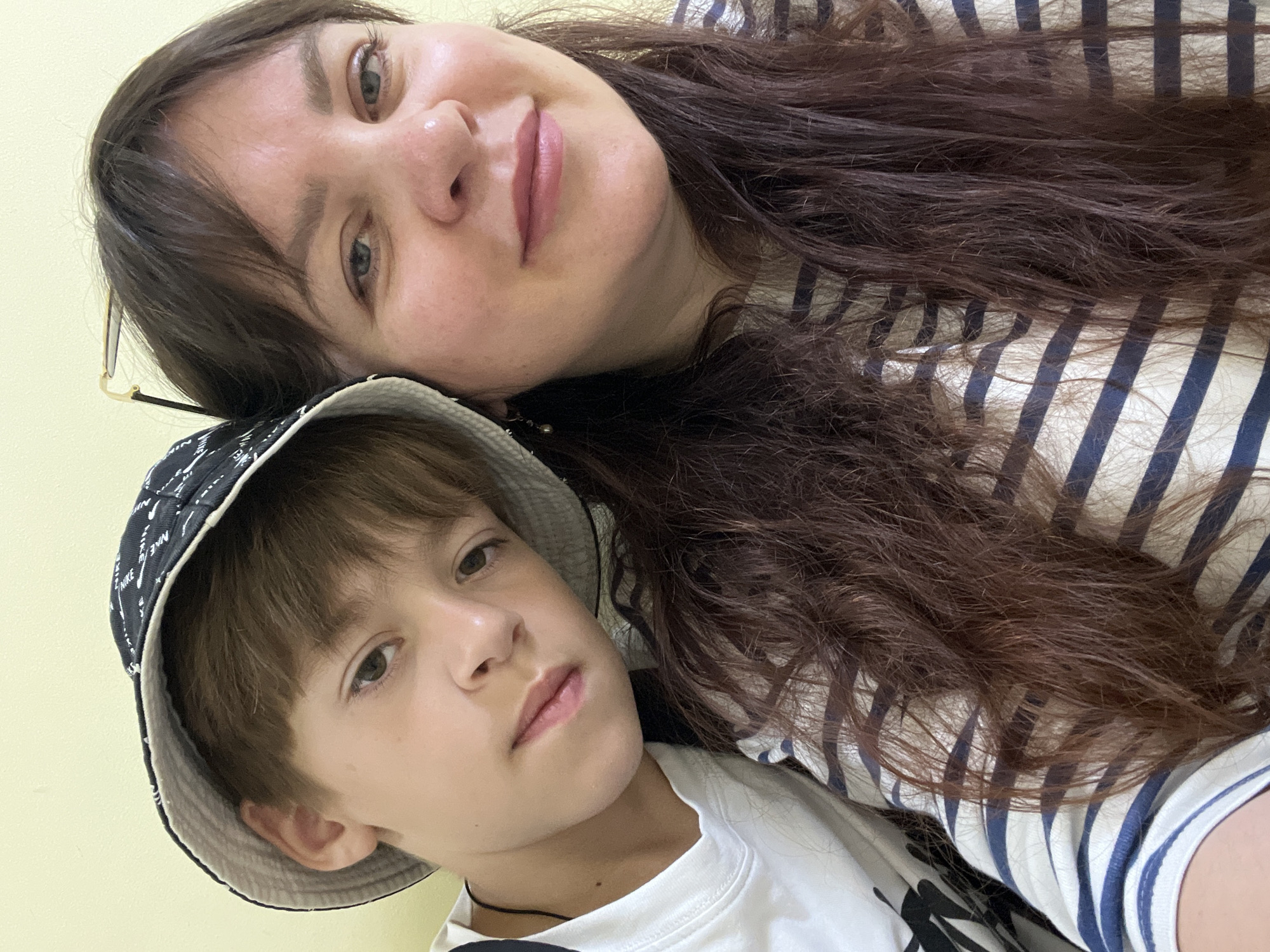 A medium shot captures a selfie of a woman and a boy in a room with a pale yellow wall. The boy, on the left, wears a white t-shirt and a dark bucket hat with a white pattern. His head is tilted, resting on the woman's shoulder. The woman, on the right, has long, dark brown hair and is wearing a striped t-shirt with navy blue and cream stripes. She is smiling and looking at the camera. She has on glasses, which are perched on top of her head. The image appears to have been taken indoors in an urban environment such as Noviy, Russia.