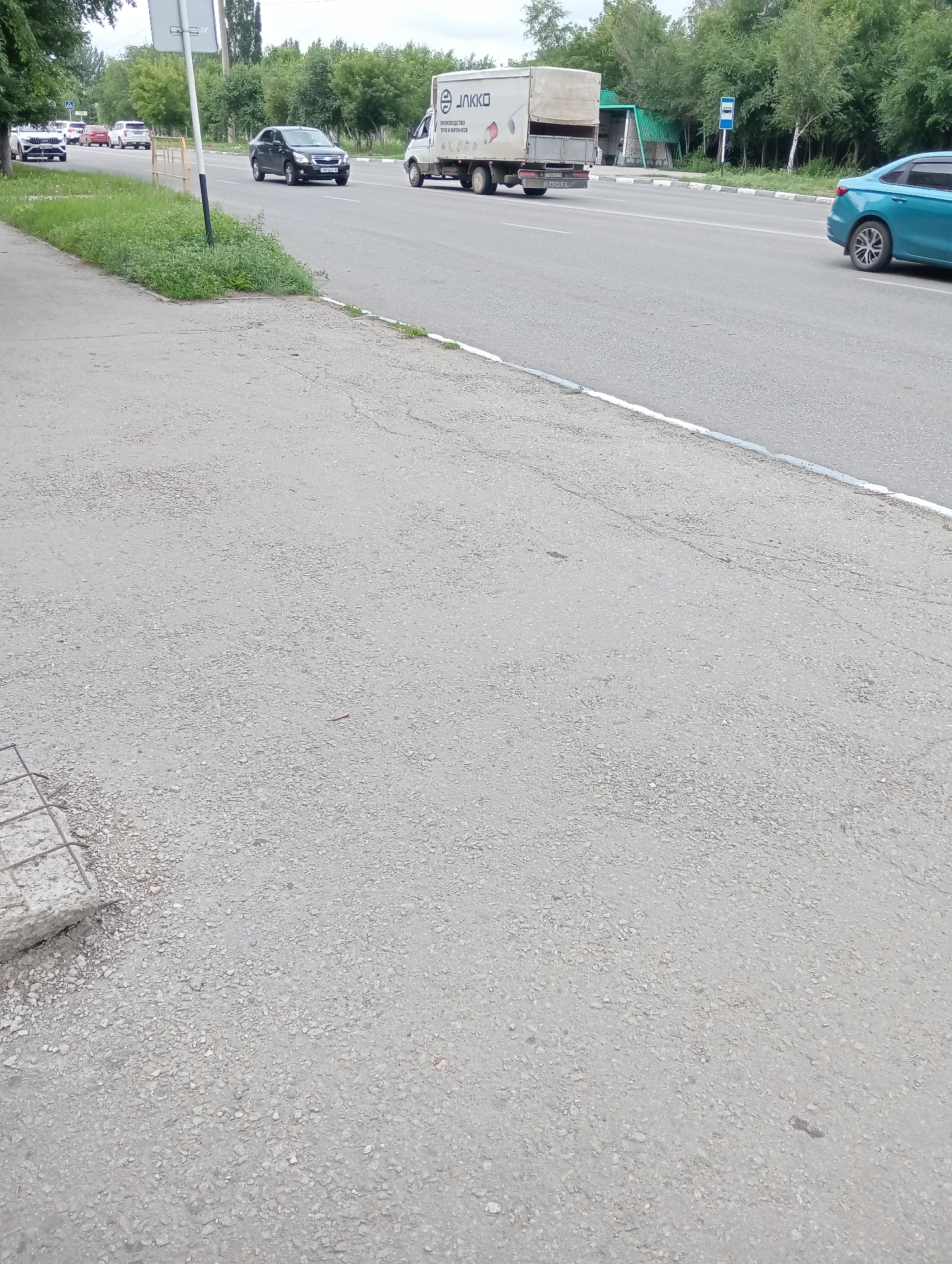 This is a medium-angle shot of a road with a sidewalk on the left. Several cars are visible on the road, including a white SUV, a black sedan, a white truck with a light-colored cargo area, and a teal sedan. The truck has text on its side that appears to be a logo with the letters "ЛЛККО" and some other Russian text below it. There is a bus stop sign visible on the right side of the road. The setting appears to be urban or suburban, with trees lining the road in the background. The sidewalk is made of asphalt and gravel, and a piece of concrete with rebar sticking out is on the lower left of the frame. The sky is overcast, suggesting it might be a cloudy day. There are no people visible in the image. The time of day appears to be daytime.