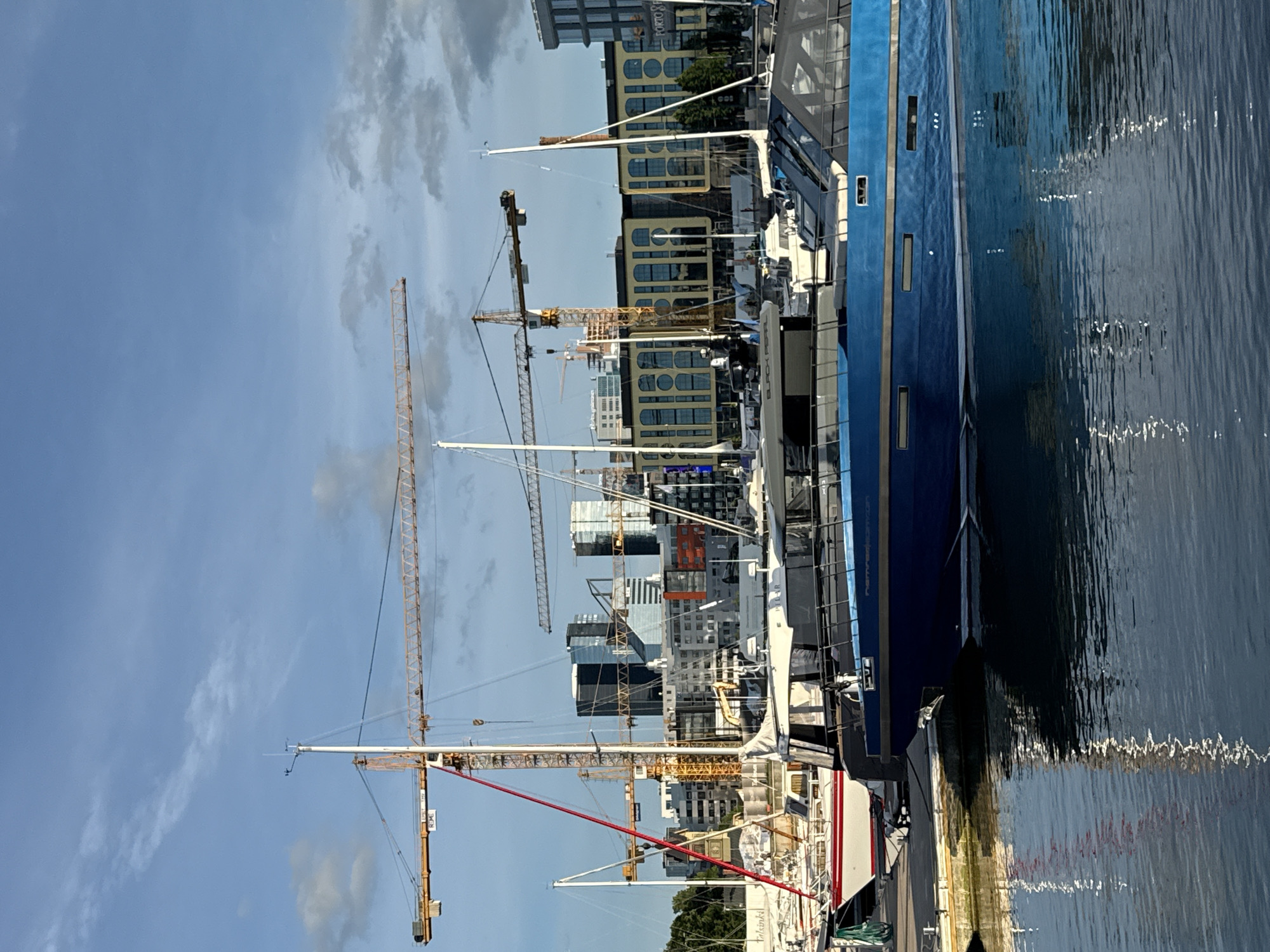 The image depicts a harbor scene in Tallinn, Estonia, likely in the Old City Harbor area, with a modern cityscape in the background. Several construction cranes are visible, suggesting ongoing development. In the foreground, a blue yacht is docked, its reflection shimmering on the calm water. Behind it, other sailboats and smaller vessels are moored. The buildings in the distance showcase a mix of modern architecture, including glass-fronted high-rises and more traditional structures with arched windows. The sky is partly cloudy, with a hint of sunlight illuminating the scene. The overall atmosphere is one of urban development and maritime activity.