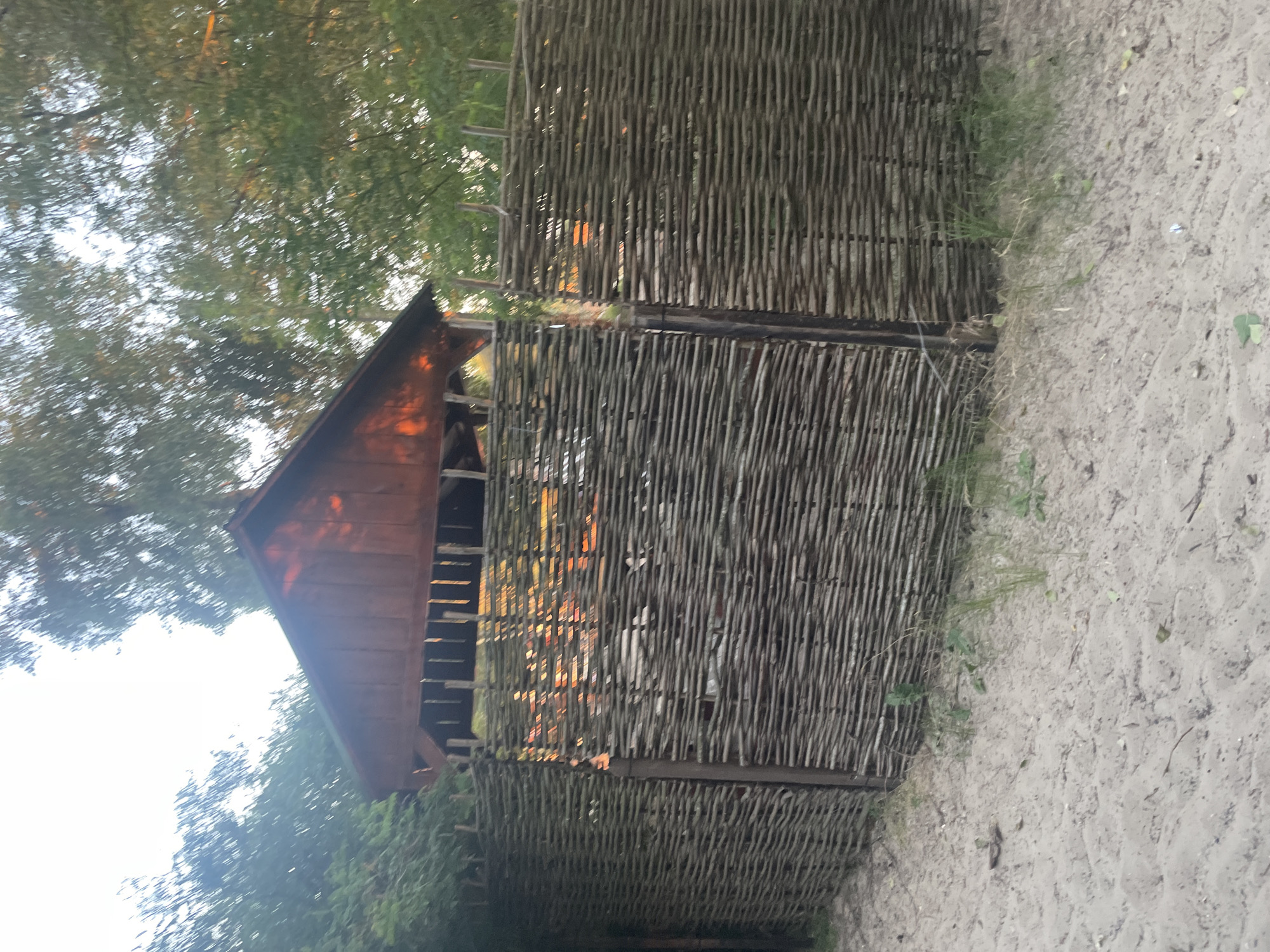 The image shows a rustic, woven twig fence in front of a wooden structure, possibly a gazebo or shed, with a peaked roof. The fence is made of tightly woven branches and stands on a sandy ground with sparse vegetation. Behind the fence, the wooden building has vertical wooden slats visible beneath its roofline, and some of the interior is illuminated by warm light, suggesting sunset or artificial lighting. Trees with green foliage are visible behind and to the sides of the building, and the sky is bright, possibly indicating daytime or late afternoon. The scene appears to be in a natural or semi-natural outdoor setting. The specific location in Kyiv, Ukraine, is not identifiable from the visual information provided.