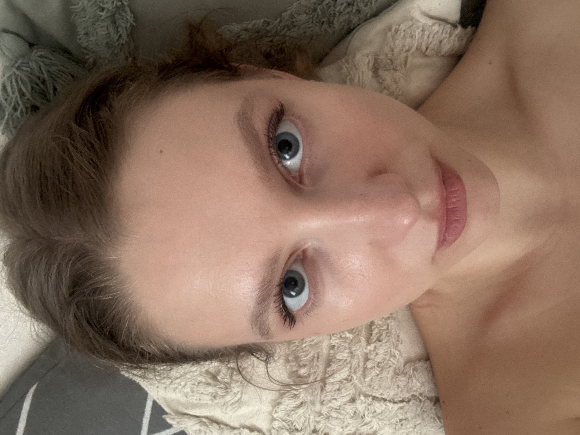 A close-up, high-angle shot shows a young woman's face and upper chest. Her eyes are wide open and directed towards the camera. She has light brown hair pulled back, revealing her forehead. Her lips are slightly parted. She is lying on a textured surface, possibly a pillow or blanket, with another textured object visible behind her head. The lighting suggests an indoor setting. No location in Izhevsk, Russia is discernible.