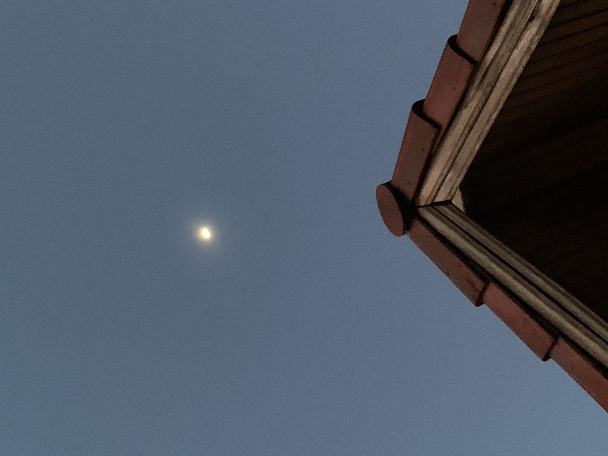 The photograph, taken at night in the city of บ้านบางรักษ์, Thailand, captures a view of the sky with a bright celestial body (likely the moon) positioned in the upper left quadrant. The sky is a uniform grey-blue. A portion of a building's roof is visible in the upper right corner. The roof is constructed with red clay tiles and wooden beams. The tiles have a curved shape, creating a stepped design. The wooden beams appear aged and are dark. The image is mostly focused on the sky and a roof.