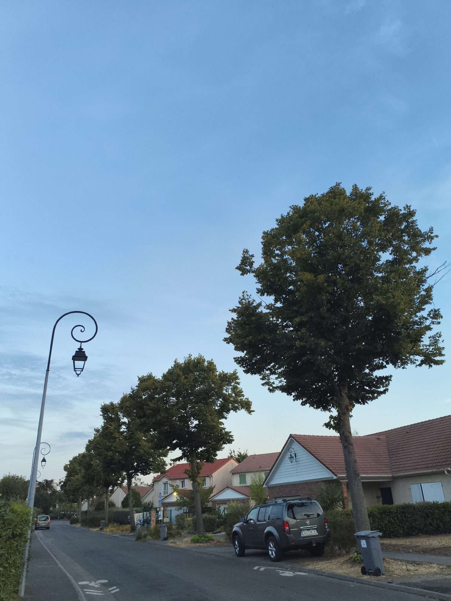 This is a street scene in the city of Carrières-sous-Poissy, France, captured during the daytime. A paved road runs through the center of the image, lined with residential houses on the right and a sidewalk and trees on the left. A dark-colored SUV is parked on the right side of the road in front of a house. Several trees with green foliage are planted along the street. A decorative lamppost with a curved arm and a lantern stands on the left side of the road. The sky is clear blue with a few wispy clouds.