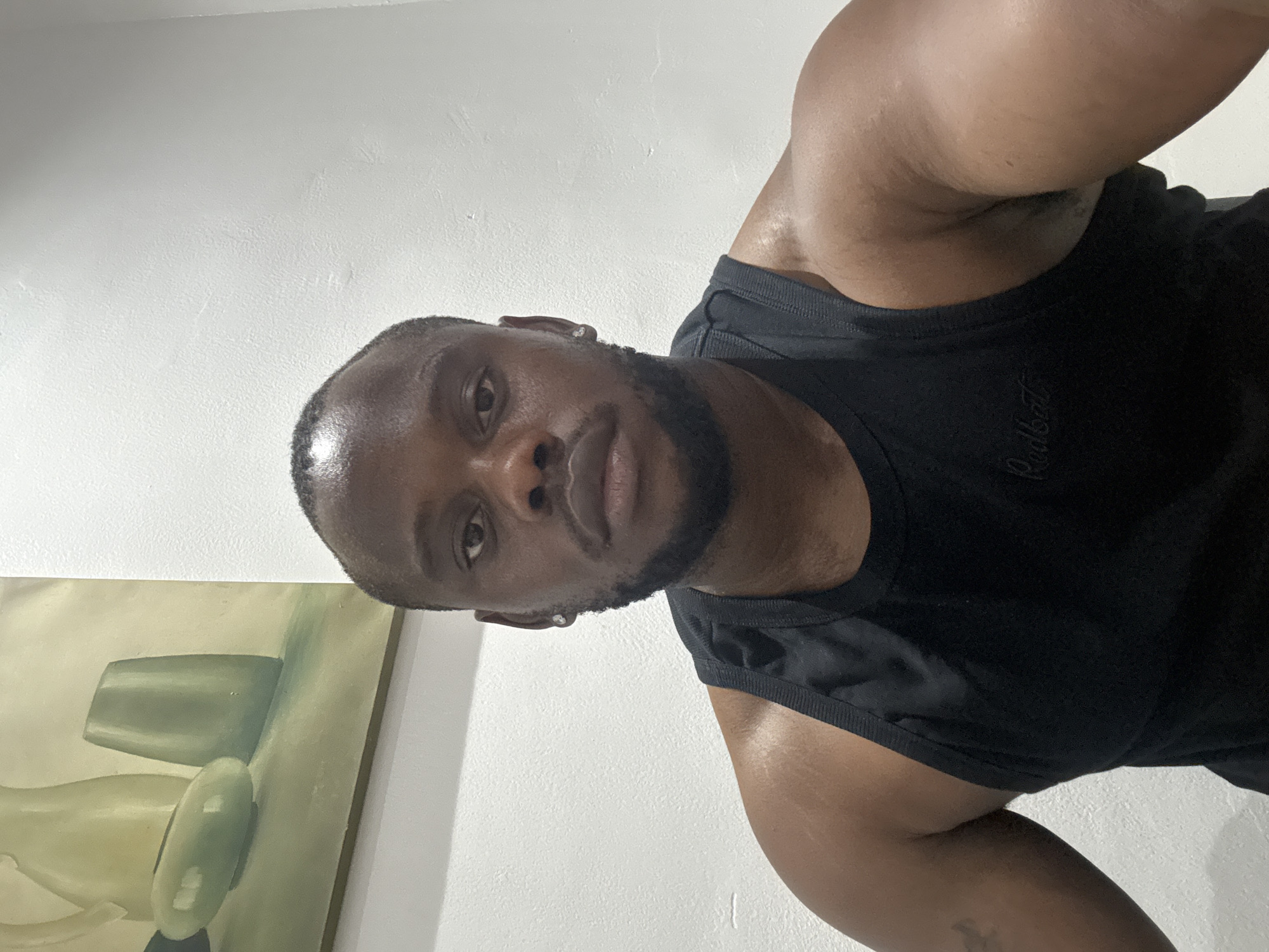 The image is a rotated selfie capturing a dark-skinned man indoors. The man has short hair, a neatly trimmed beard, and is wearing a black ribbed tank top. A small, silver-colored stud earring is visible in his left ear. He looks directly at the camera with a calm expression. His muscular arms and upper torso are prominent, and his skin has a slight sheen, possibly from sweat.
The background consists of a plain, light-colored (likely white or off-white) wall. To the left of the man, a framed painting is visible on the wall, featuring abstract or still-life objects in muted green, beige, and grey tones. The overall lighting suggests an indoor setting, possibly with artificial light.
Visible text on the man's tank top, near the chest, is the word "Redbat" in a subtle, embroidered script font. The image is rotated approximately 90 degrees counter-clockwise, suggesting it was taken from an unconventional angle, perhaps while the subject was lying down. The location context is Iddo Tudunwada, Nigeria.