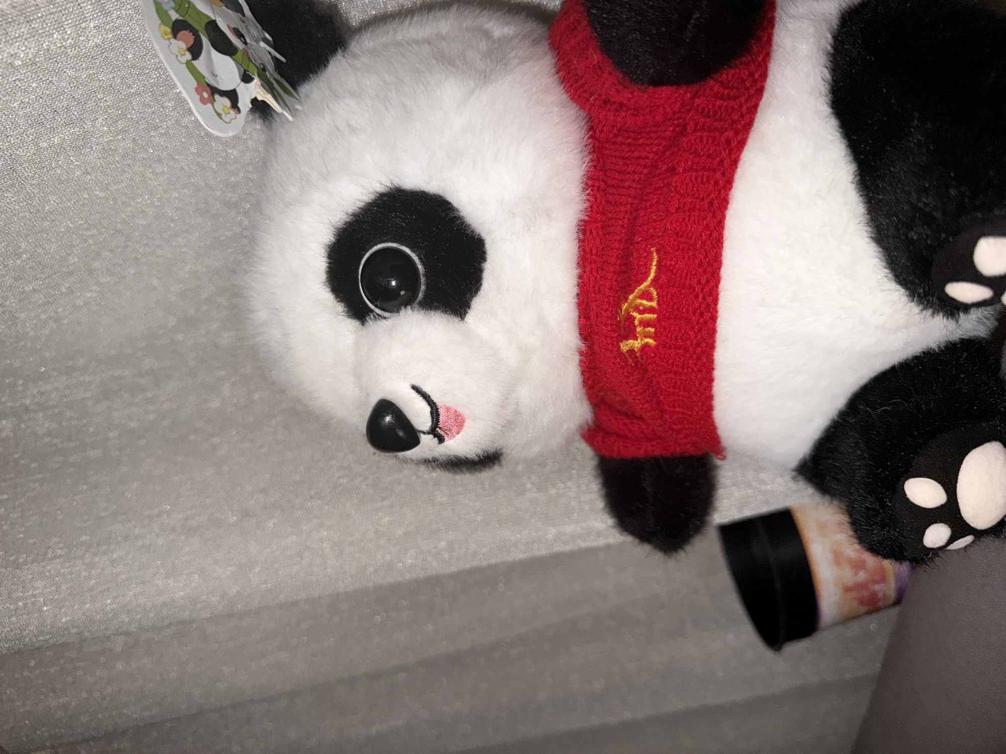 A stuffed panda toy wearing a red knitted scarf lies on a textured gray surface. The panda's head is angled towards the left, revealing a black eye patch, a black nose, and a pink mouth. A black and white tag is visible near the top left corner. Part of a label with faded colors and a black ring is visible near the panda's paw in the lower right corner. There is no evidence to suggest that the image was taken in Sofia, Bulgaria.